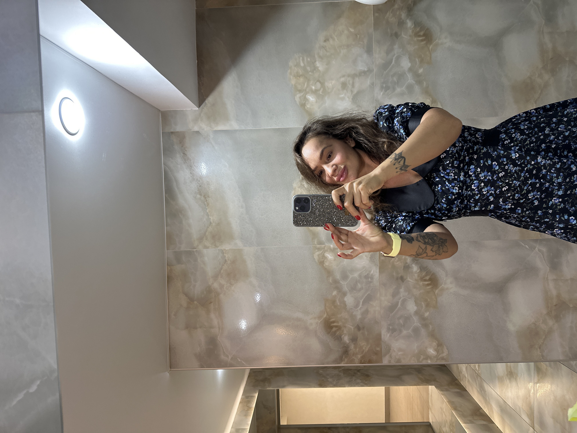 A woman is taking a selfie in a tiled bathroom. She is wearing a dark floral dress with a Peter Pan collar and has wavy brown hair. Her left arm, visible in the frame, displays tattoos and a yellow band on her wrist. She is holding a smartphone with a sparkly case. The bathroom walls are covered in large, beige marble-patterned tiles. The ceiling is white with a circular light fixture. To the left, a doorway leads into another area with beige tiling and a light. The scene appears to be an indoor domestic or public restroom. There is no indication of the city of Duvankul', Russia.