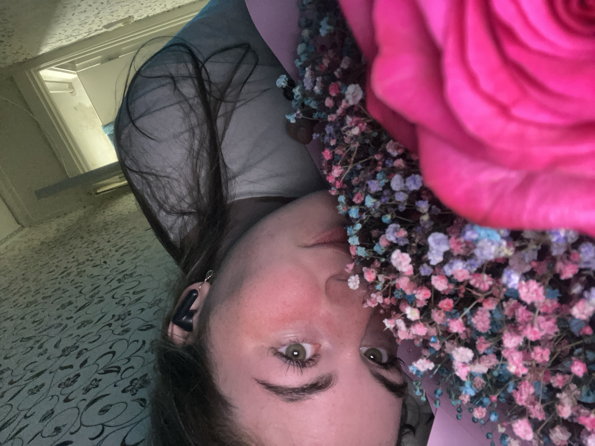 A young woman is seen in a close-up portrait, holding a bouquet of flowers, including a large pink rose and smaller, colorful baby's breath. She has long, dark hair, fair skin, and green eyes. An earbud is visible in her left ear. The background shows a patterned surface, possibly a bedspread or carpet, and glimpses of a ceiling and interior doorway. The image does not provide any information to confirm the location of the photograph being taken.