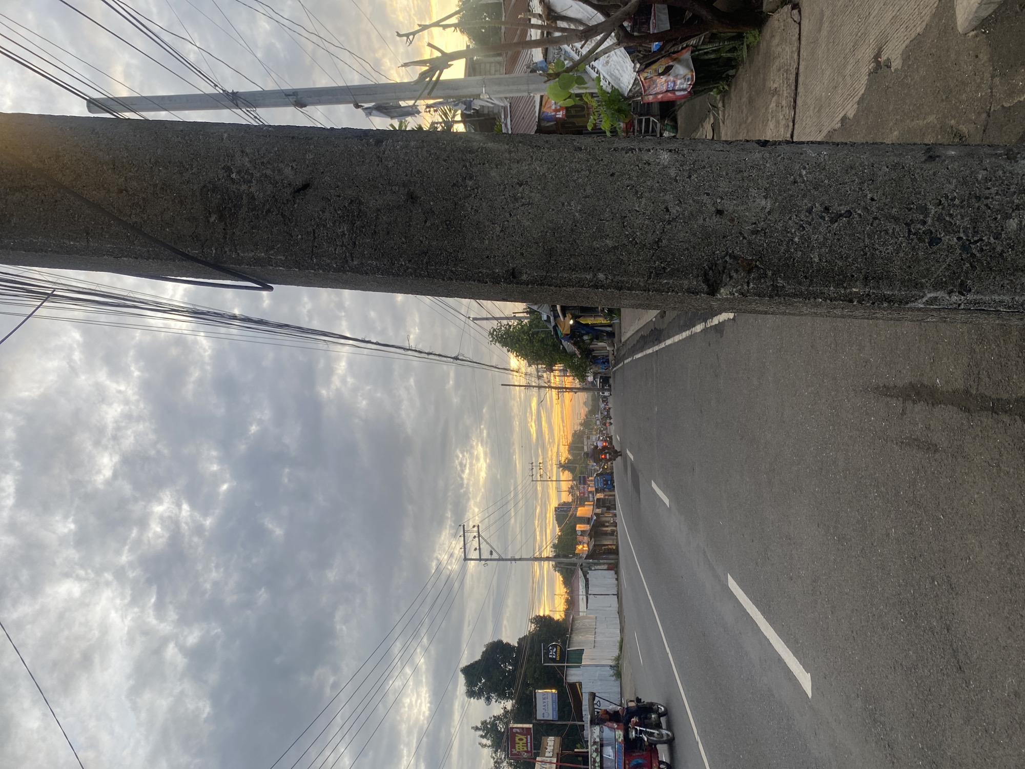 The image shows a street scene in Davao City, Philippines, at sunrise or sunset. A concrete utility pole dominates the foreground on the right, with a network of overhead wires crisscrossing the sky. The road, which appears to be asphalt, has white lane markings. On the left side of the road, there are buildings with signs, suggesting a commercial area. A tricycle, a common mode of transport in the Philippines, is seen on the road. Several other vehicles, including motorcycles, are visible further down the street. The sky is filled with clouds, with hints of orange and yellow light indicating the sun's position. The overall impression is of a busy street during transitional light conditions in a tropical urban environment.