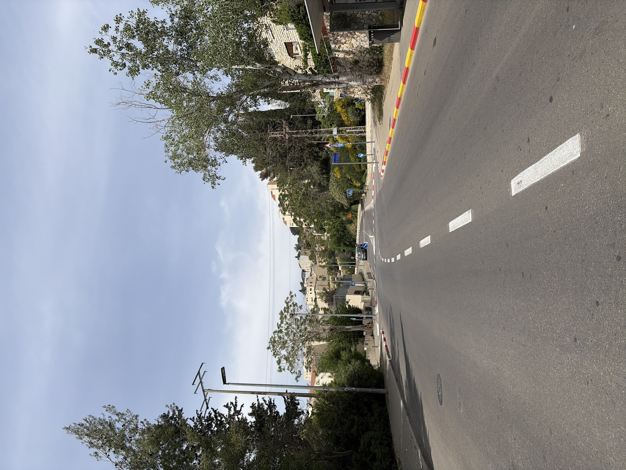 The image depicts a street view in an unidentified urban location. The street is paved with asphalt, marked with white lane dividers and a red and yellow curb line. The road ascends slightly into the distance, bordered by buildings, trees, and vegetation. Overhead utility lines are present, supported by poles, and a blue sky with scattered clouds is visible. There are several street signs and a vehicle present further up the road. The scene is well-lit, suggesting daytime.