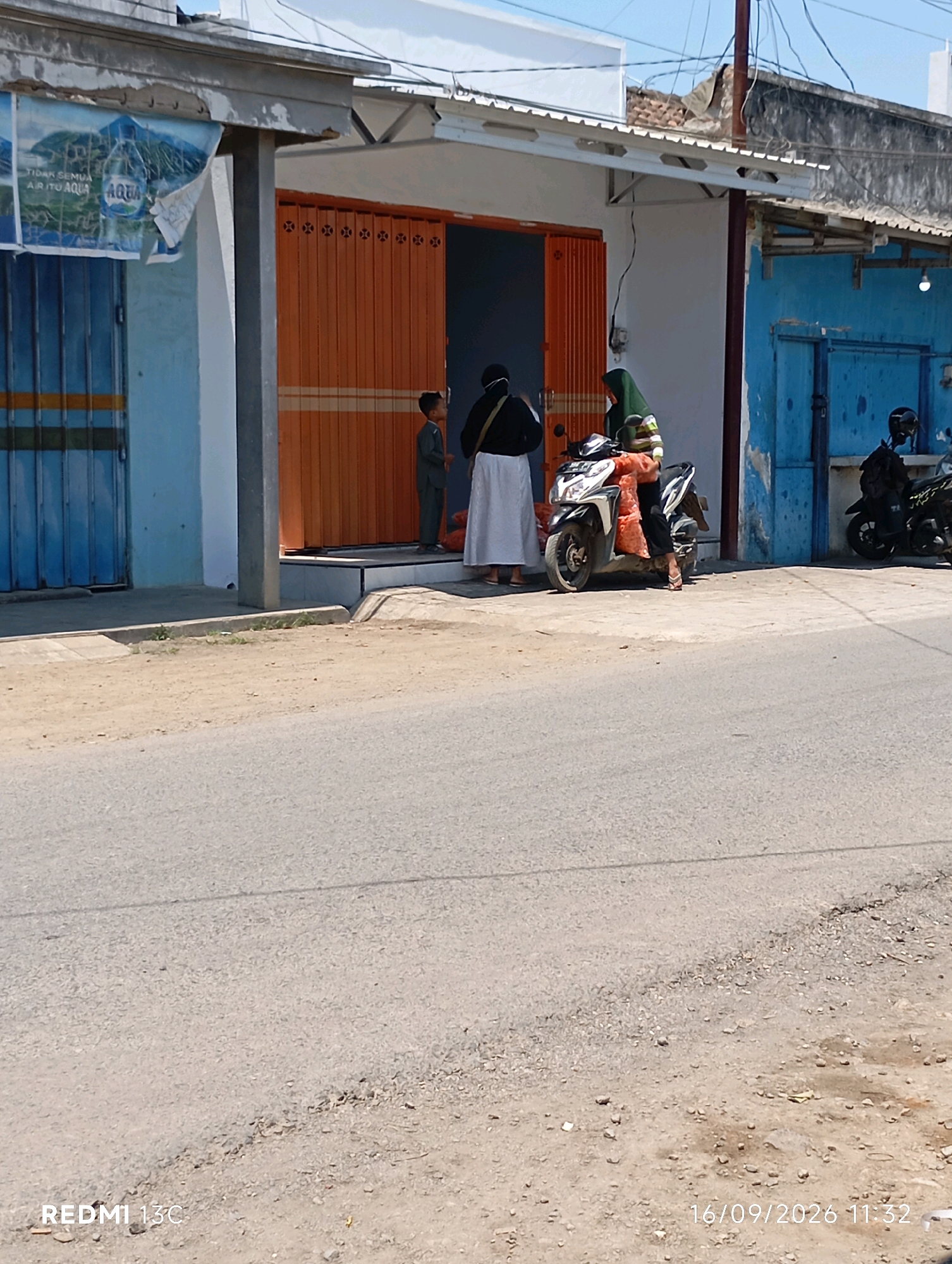 The image captures a street scene in Wuluhan, Indonesia, on September 16, 2026, at 11:32 AM. In the foreground, a paved road transitions to a sandy shoulder with a concrete curb. A building with orange roll-up doors and blue shutters occupies the mid-ground. Two individuals are visible near the orange doors. One, a child in a gray uniform, stands to the left. To the right, an adult wearing a green hijab and a striped shirt is seated on a motorcycle, which has a large orange bag on the back. Another motorcycle with a rider is parked to the right of the building. The building's facade appears somewhat weathered, with peeling paint on the blue shutters. A banner with the AQUA logo is partially visible on the left side of the building. The sky is clear and blue.