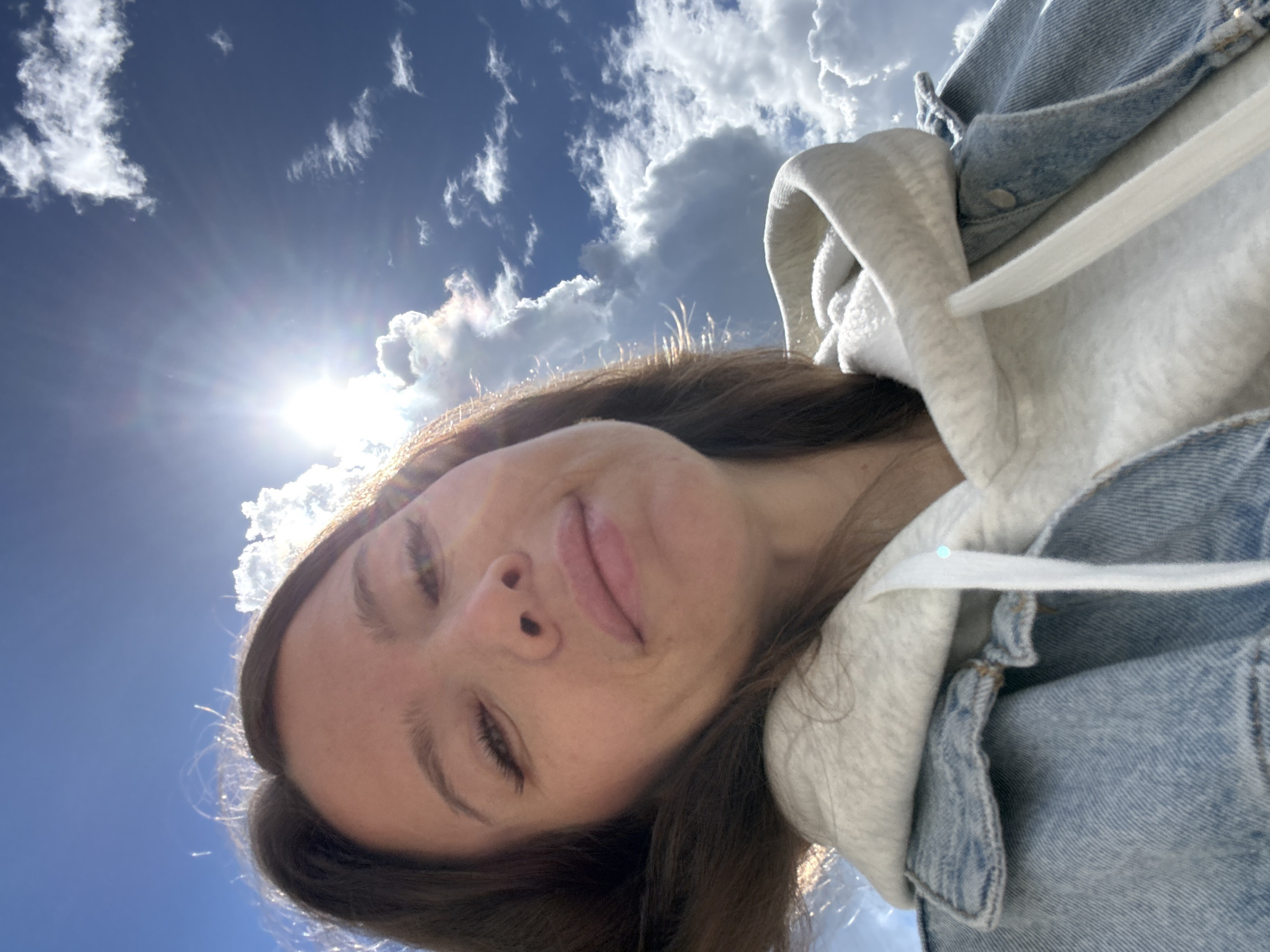A selfie, likely taken outdoors given the sky background. The sky is blue with white clouds and the sun is visible, creating a bright glare. A person with brown hair and closed eyes, wearing a white hoodie under a light blue denim jacket, is in the foreground.
There is no indication of location in the city of Moscow, Russia.