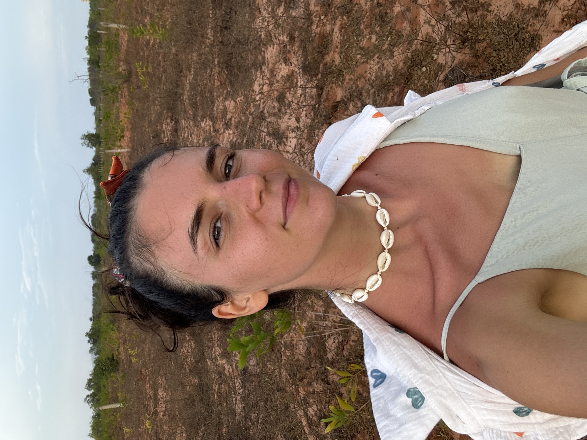 A woman is in an outdoor location in Phu Thuy Ward, Vietnam. The background consists of dry, reddish-brown earth with scattered vegetation, including small trees. The sky above is a pale, hazy color. The woman is wearing a light green top, with a white patterned shirt draped over her shoulders. She has a necklace made of shells. Her dark hair is pulled back, and she has an ornament in her hair. She is looking directly at the camera with a slight smile.