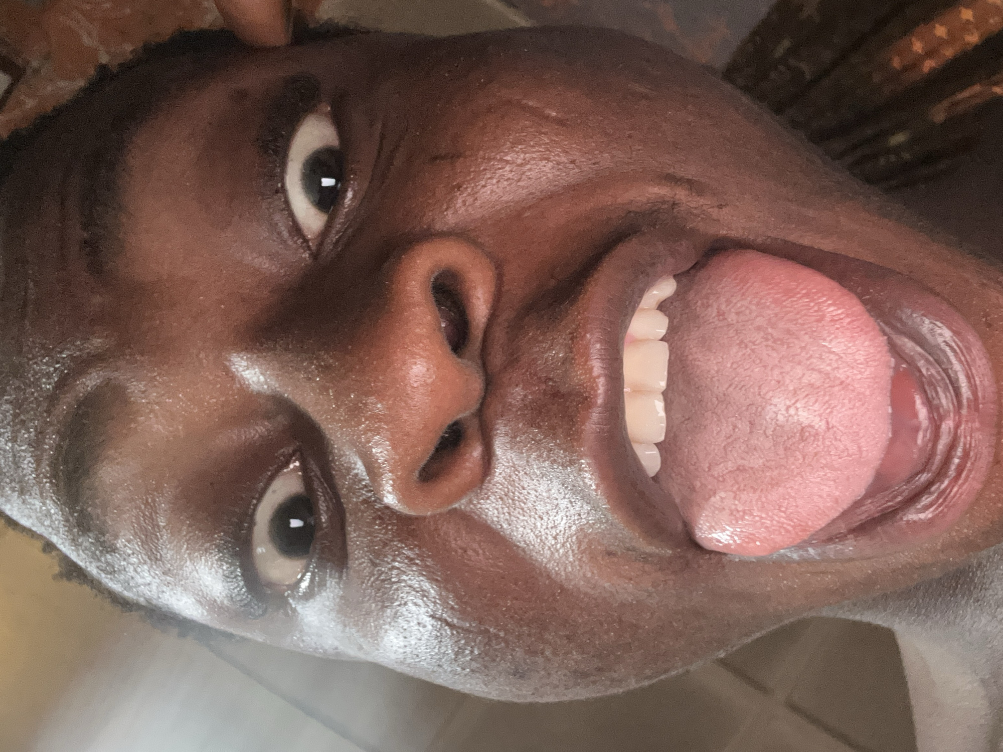 A close-up, upward-angled shot captures a person with dark skin. The subject's mouth is open, revealing their tongue extended outwards and their top teeth. Their eyes are wide, and their face appears shiny. The background is out of focus.