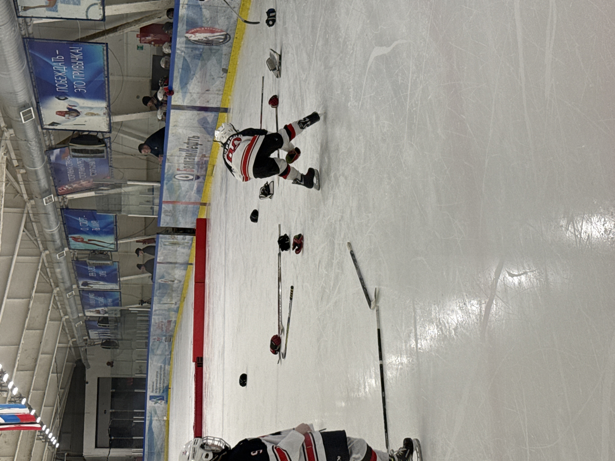 Here is a detailed description of the image:
The image depicts a scene on an ice rink, likely during or after a hockey practice or game. Several hockey sticks are scattered on the ice, along with gloves and a puck. Two hockey players are visible. One player is located near the top center of the frame, and the other is closer to the bottom. Both are wearing hockey equipment, including helmets, pads, and skates. The player near the top center seems to be in a dynamic pose.
The rink's boards are visible along the left side of the frame, marked with red and yellow lines. Above the boards are advertising banners, including one that reads "ПОБЕЖДАТЬ - ЭТО ПРИВЫЧКА!" (To win is a habit!) and "Белкам Нефть". The banner also indicates that the rink is in Izhevsk, Russia, as it is written in Russian. Spectators can be seen behind the boards in the distance. The environment suggests an indoor hockey arena or ice rink setting.