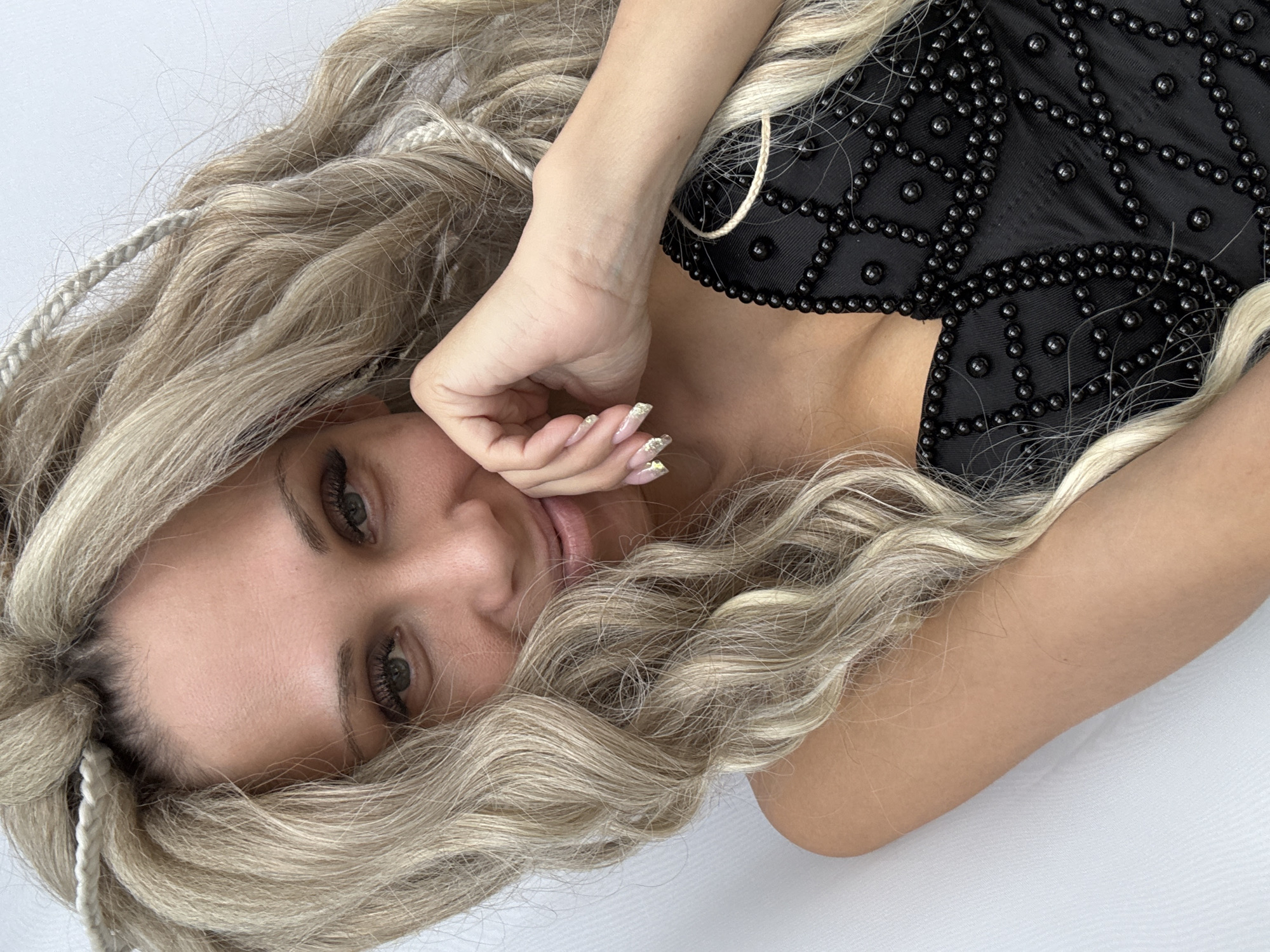 A woman with blonde hair and braids is lying down on a white surface. She is wearing a black top adorned with beads. Her right hand is near her face, and she is looking directly at the viewer. The location is unspecified and there is no indication the location is Rusanovo, Russia.