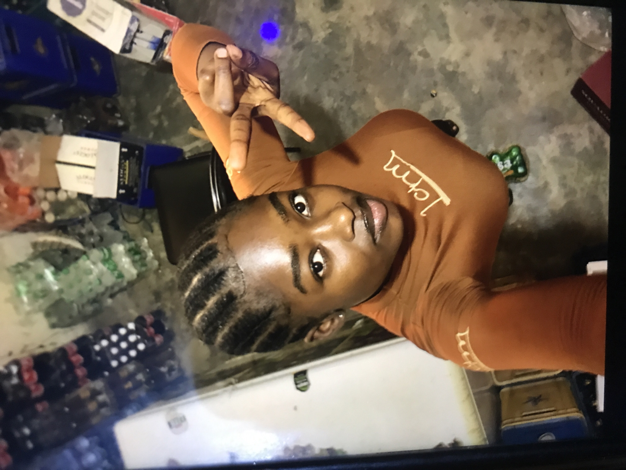 A woman with cornrow braids is depicted from a top-down perspective. She is wearing an orange long-sleeved shirt with "I Am" printed in white and is holding up a peace sign with her right hand. The background is cluttered with various objects, including blue plastic crates, boxes, and other miscellaneous items. The setting appears to be indoors, likely a storage or utility area. This scene is within the city of New Stadium, Nigeria.