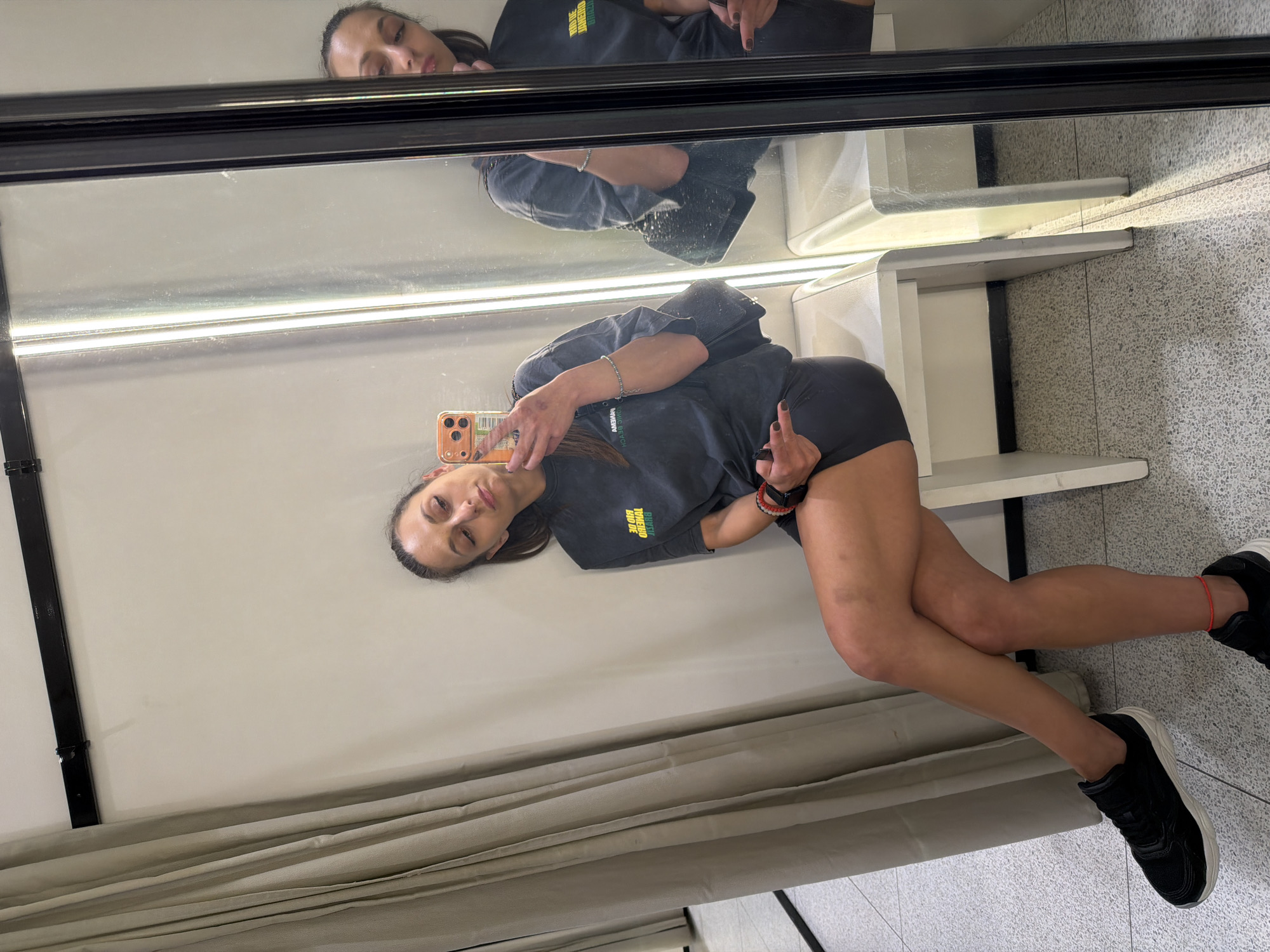 A woman with dark hair tied back is sitting in a fitting room. She is wearing a dark grey t-shirt with yellow lettering that reads "RIO DE JANEIRO BRAZIL" and dark grey shorts. She is holding a phone in her right hand and taking a selfie in a mirror. She has on black sneakers with white soles and red string tied around her left ankle. A black purse with a chain strap is visible over her left shoulder, and she is wearing a bracelet on her left wrist and a red bracelet on her right wrist. The fitting room has beige curtains on the left and a white bench she is sitting on. The floor is a speckled grey. The image is taken in Plovdiv, Bulgaria.