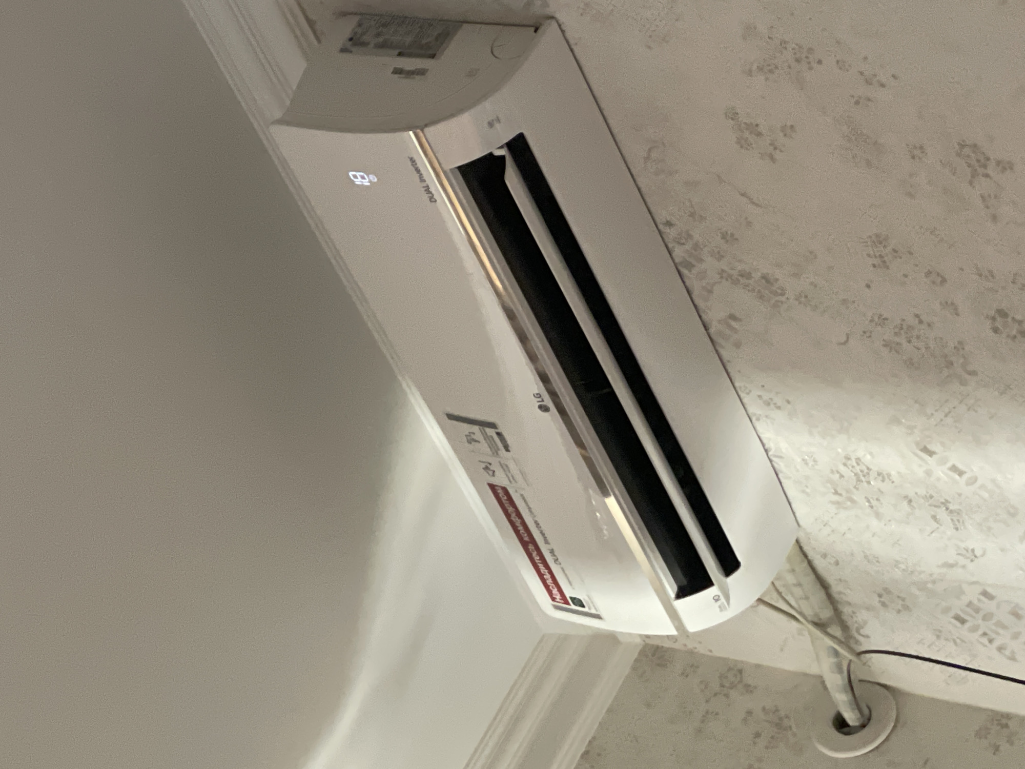 The image shows a white LG air conditioner mounted high on a wall. The air conditioner has a digital display showing "18". A red sticker with Russian text is visible on the front of the unit, and the "LG" logo is also present. Below the air conditioner, a white pipe runs horizontally along the wall, secured by a circular bracket. The wall has a textured pattern. The ceiling above the air conditioner is white with decorative molding. The image appears to have been taken indoors, likely in a residential or commercial space. The location is identified as Tashkent, Uzbekistan.