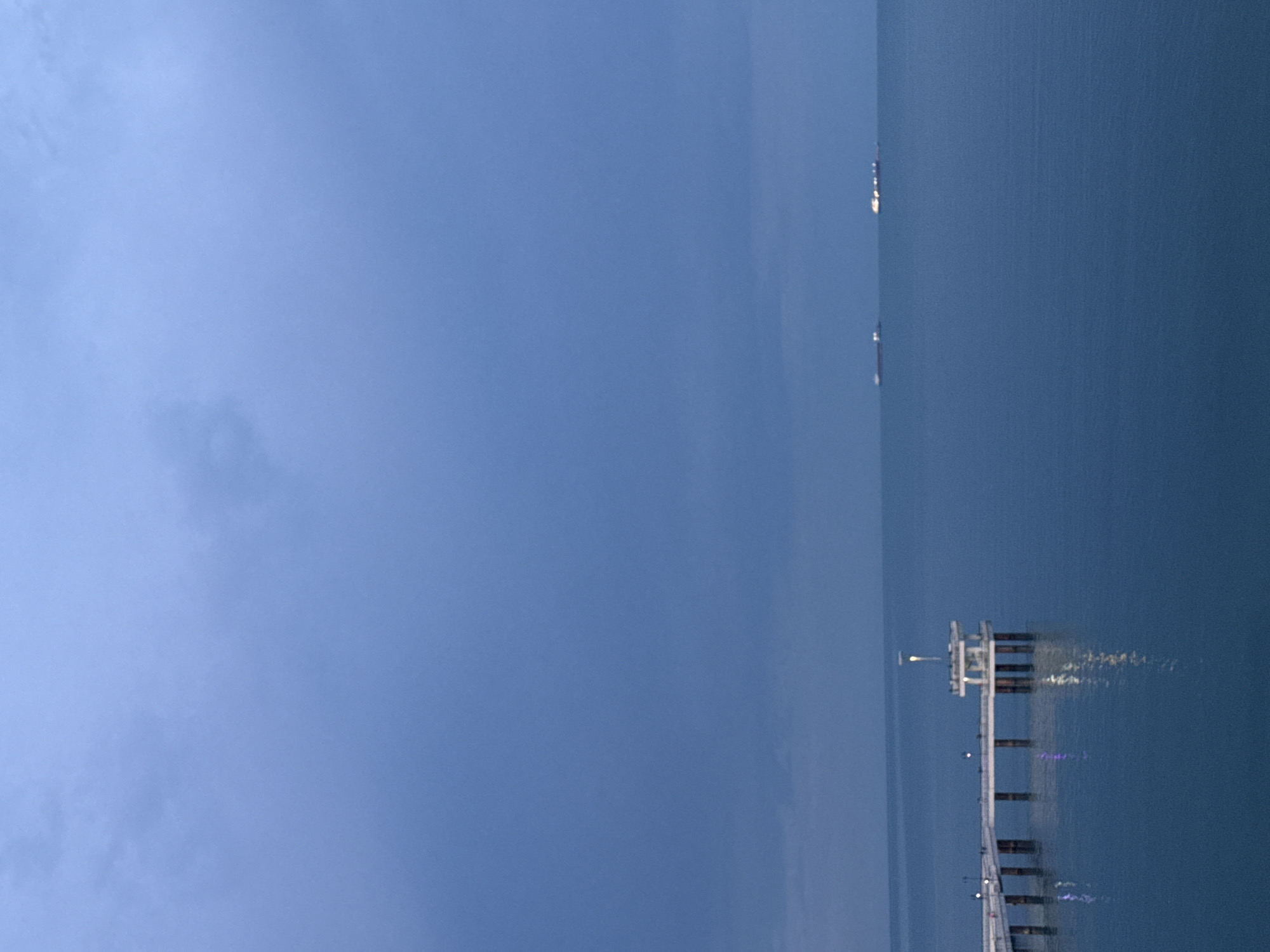 The image shows a view of the sea at Burgas, Bulgaria, taken at dusk. A pier extends into the water from the right. The sky is mostly blue with some cloud cover. Two ships are visible in the distance, aligned along the horizon in the upper-right. The pier is well-lit with numerous lights, casting reflections on the water's surface.