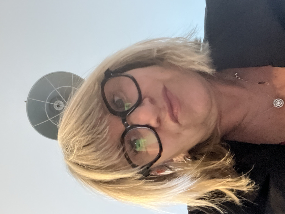 A woman with blonde, shoulder-length hair and black-rimmed glasses poses close to the camera, resulting in a low-angle perspective. Part of a modern lamp fixture is visible in the upper left corner. She's wearing a black top and a delicate necklace with a circular pendant. Her expression is neutral, and the background appears to be a light-colored wall. The image captures a casual, personal moment.