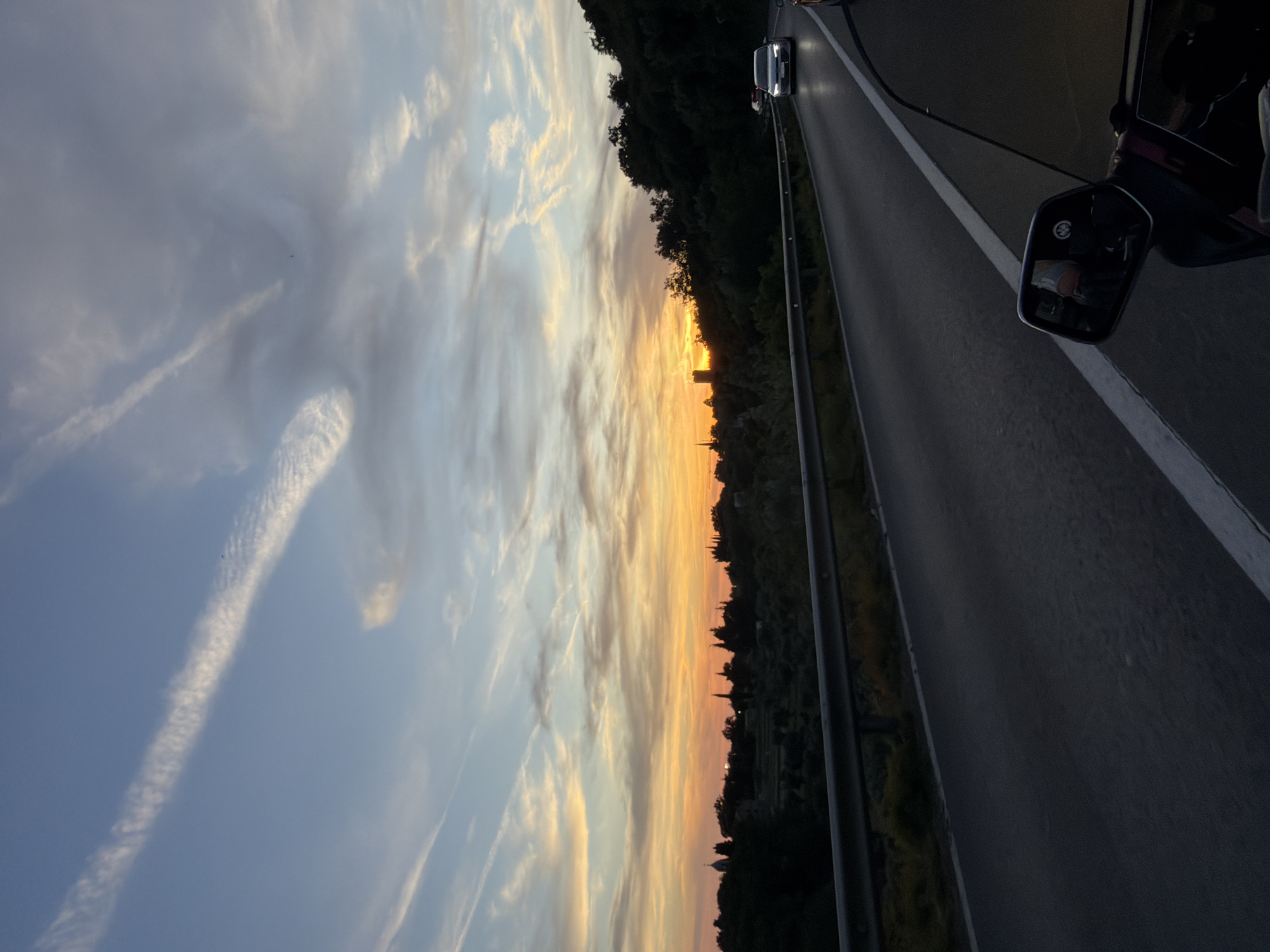 This is a photograph taken at dusk from a vehicle traveling on a road in Gornji Katun, Montenegro. The sky is painted with the warm hues of sunset, with oranges, yellows, and blues blended with scattered wispy clouds. In the distance, silhouetted against the vibrant sky, are the shapes of trees and buildings, including what appears to be a church steeple and possibly a tower. The road ahead is paved, with a white line marking the lane. A metal guardrail separates the road from the embankment. Several cars are visible on the road, moving in the same direction. In the foreground, a side-view mirror of the vehicle is visible, reflecting a passenger. The surrounding landscape is hilly and covered with dense, dark green foliage, suggesting a rural or semi-rural setting.