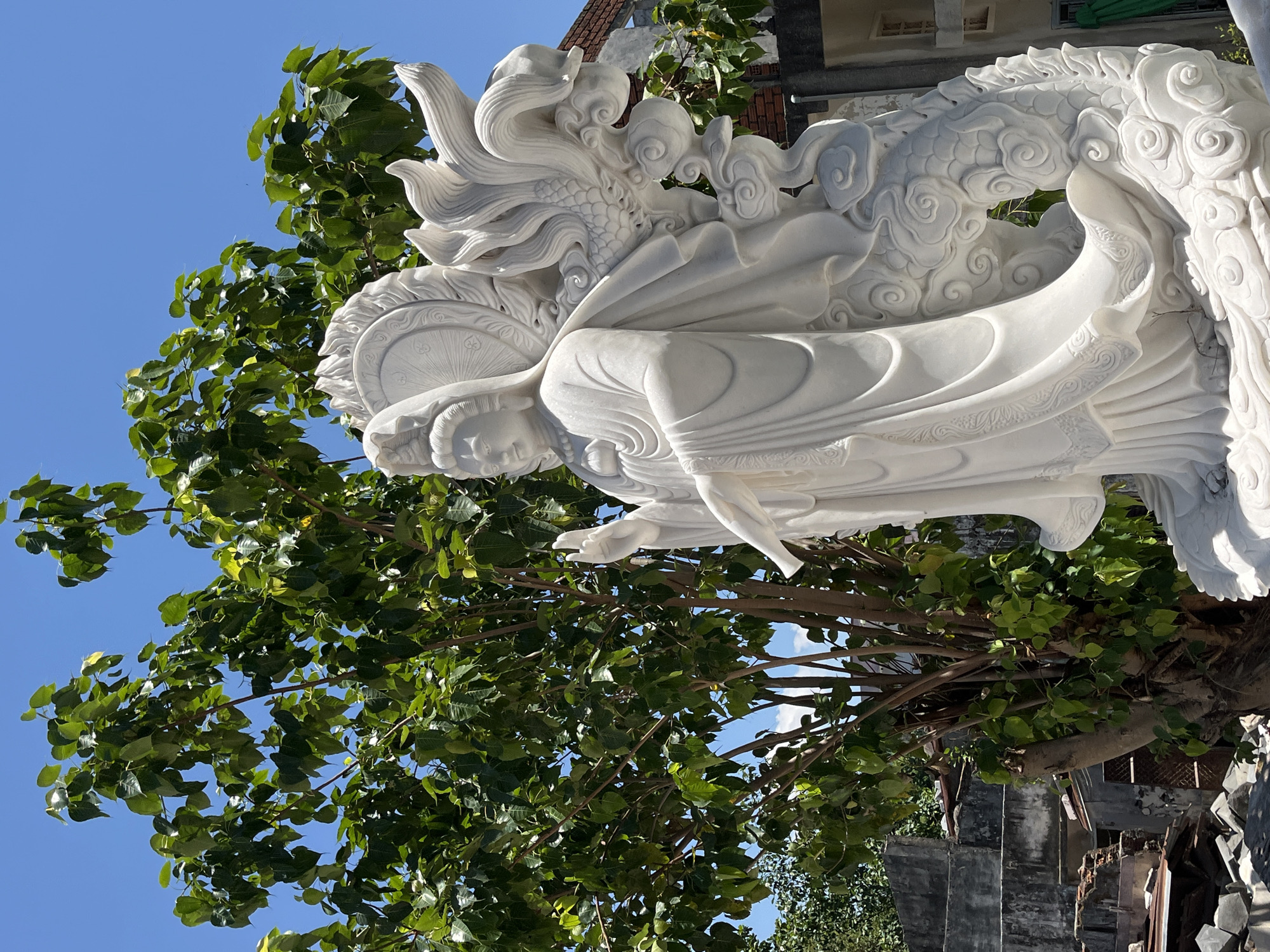 The image depicts a white marble sculpture of a woman with a dragon, set against a backdrop of a tree with green leaves and a clear blue sky. The sculpture appears to be outdoors, possibly in a garden or park setting. The woman is depicted with flowing robes and a serene expression. The dragon coils around her, adding a mythological element to the sculpture.
Based on visual details and known landmarks, the statue is likely located in Đà Nẵng, Vietnam. Đà Nẵng is known for its marble sculptures, particularly around the Marble Mountains area.
