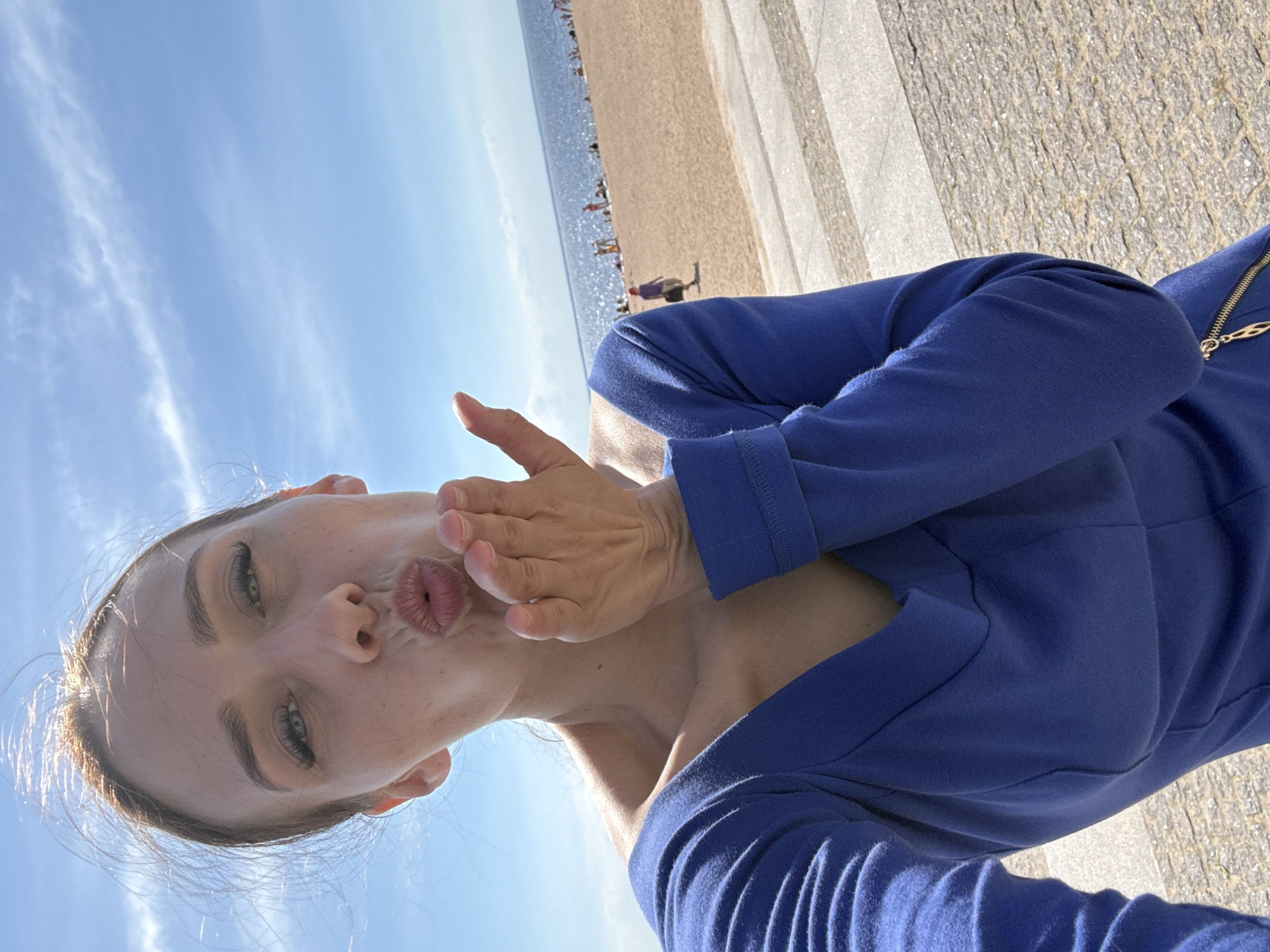 A woman in a blue dress is taking a selfie on a sunny day at the beach. Her hair is blowing in the wind. She is puckering her lips as if to blow a kiss. In the background, the ocean and a sandy beach with people are visible. This appears to be a scene from a beach in Saint Petersburg, Russia, possibly on the Gulf of Finland.