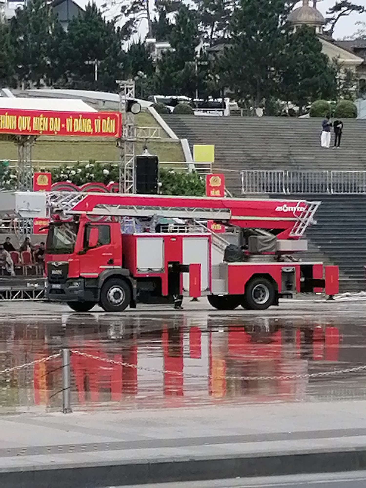 The image shows a red fire truck with a ladder parked on a wet, reflective surface, likely a plaza or street in Da Lat, Vietnam. The fire truck is a modern vehicle, possibly a MAN model, with its ladder extended. In the background, there are tiered stone stairs leading up to buildings with domed roofs, surrounded by lush green trees. A red banner with Vietnamese text is visible above and to the left of the fire truck, with the text reading "CHÍNH QUY, HIỆN ĐẠI - VÌ DÂN, VÌ ĐẤT". Several people are seen on the stairs in the distance, and a few more are seated in chairs to the left of the fire truck. The overall scene suggests an outdoor event or demonstration in Da Lat, a city known for its French colonial architecture and mountainous terrain. The wet surface indicates recent rain or a cleaning.