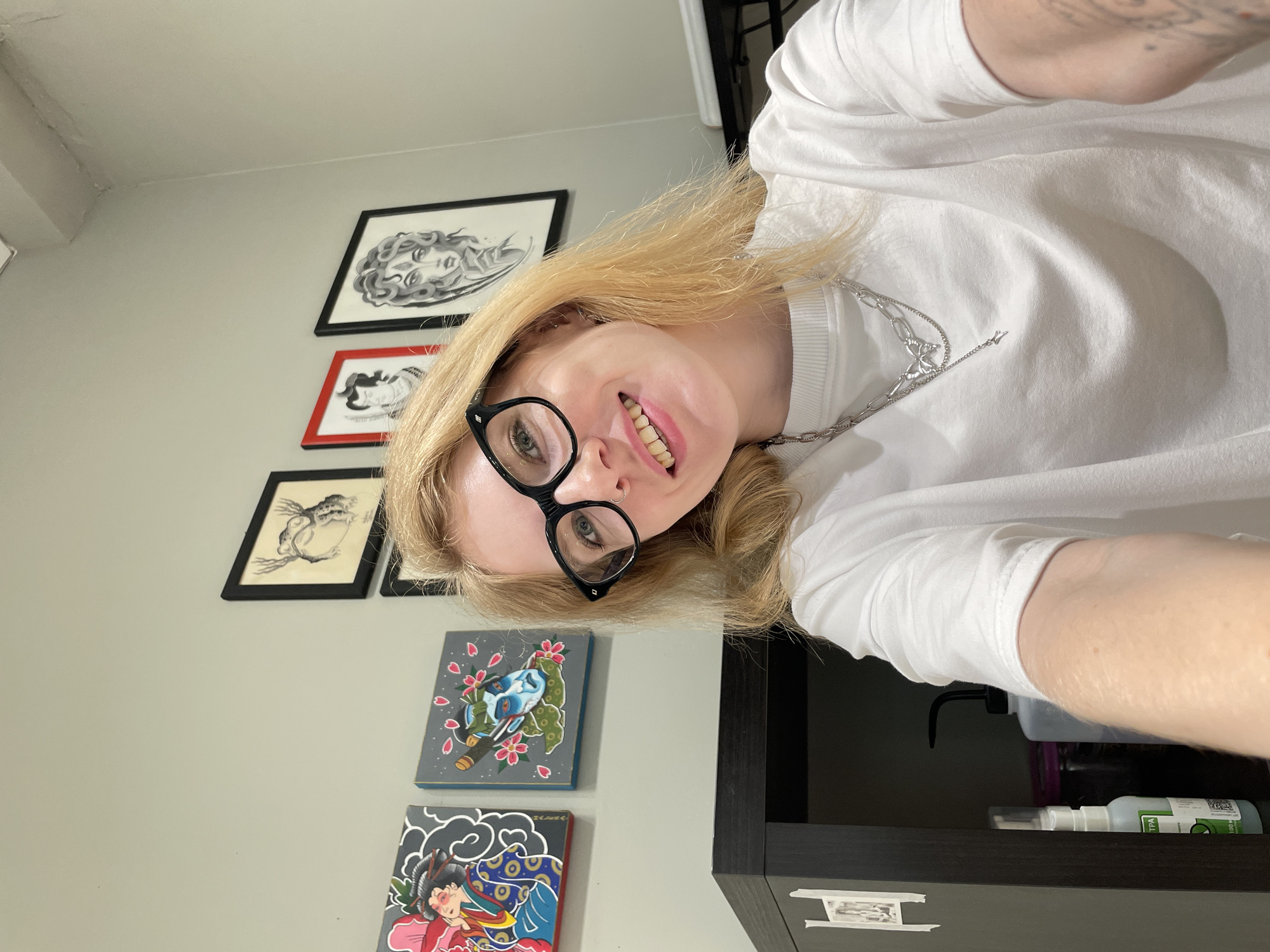 A young blonde woman with blue eyes and glasses is taking a selfie. She is wearing a white t-shirt and a silver necklace with a butterfly pendant. In the background, there are several framed artworks on a light green wall. To the left, on a dark shelf, are various bottles and containers, including a spray bottle. The scene appears to be an indoor studio or workspace, possibly a tattoo parlor, given the artwork. No specific location in Izhevsk, Russia can be determined from the image.
