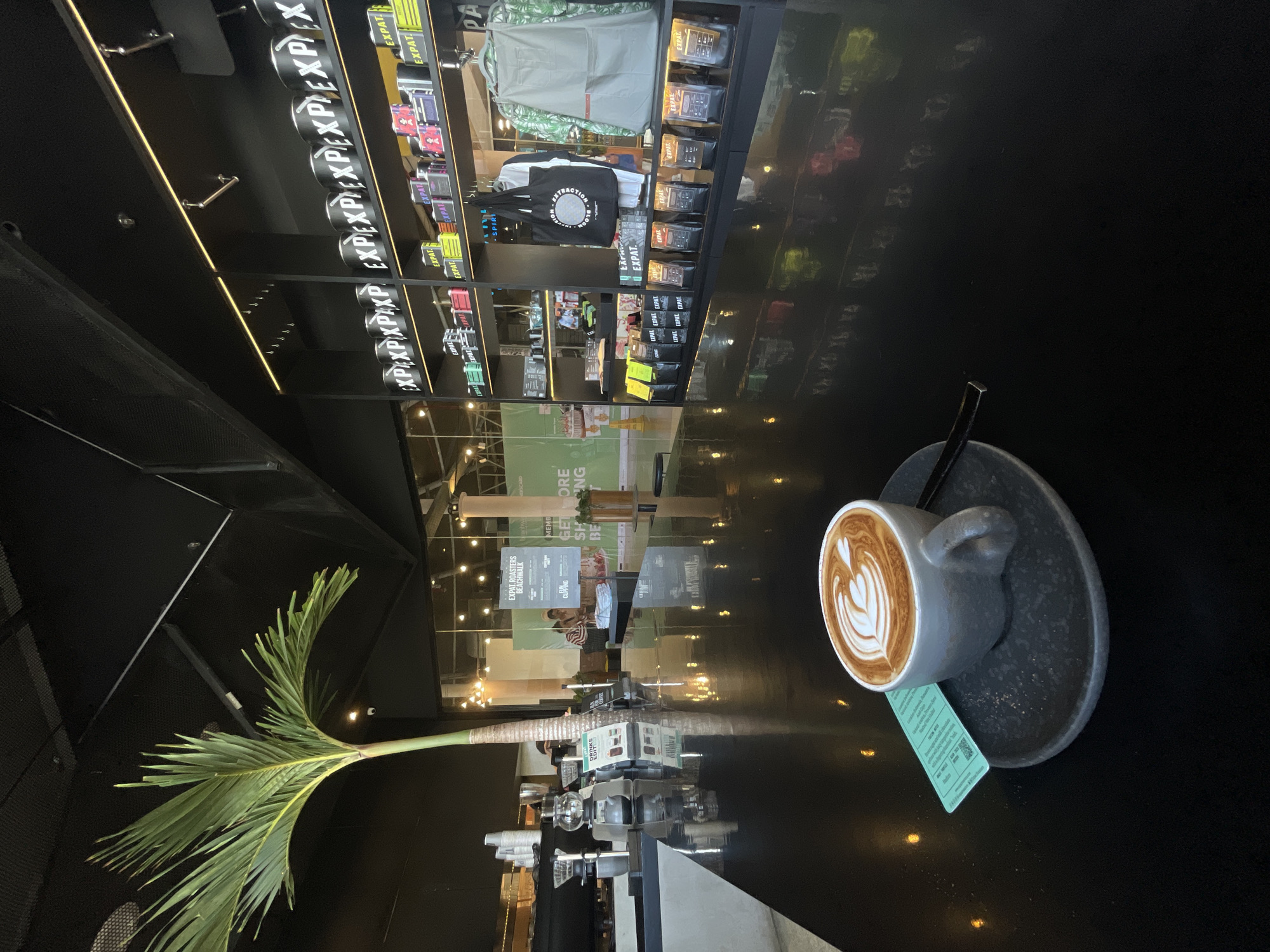The image displays a modern, chic cafe interior, likely "EXPAT ROASTERS BEACHWALK" in Kuta, Indonesia, as indicated by a prominent sign and the presence of a palm tree indoors. In the foreground, a dark grey ceramic coffee cup, adorned with intricate latte art featuring a heart-leaf design, sits on a matching saucer on a highly reflective black table. A small green tag is tucked beneath the cup.
To the left, a tall, slender palm tree with lush green fronds adds a distinct tropical ambiance to the contemporary setting. The background features extensive dark shelving units showcasing various products. Many items, including numerous coffee bags in different packaging colors (black, yellow, red, green, purple), are labeled "EXPAT" and "EXPAT ROASTERS." Merchandise like a black tote bag with "EXTRACTION ROAST" printed on it and a green-patterned apron are also displayed. The shelves are illuminated by subtle strip lighting.
Further into the background, a counter area with coffee-making equipment is visible. Patrons are seated at tables or counters, appearing relaxed. The reflective surfaces throughout the cafe cast mirror images of the lights, shelves, and distant figures, contributing to the sleek aesthetic. Visible text includes "DRINKS," "EXPAT ROASTERS BEACHWALK," "EXPAT" (repeatedly), "EXTRACTION ROAST," and parts of a sign reading "MEMB" for a membership program. The scene suggests a calm and inviting atmosphere typical of a specialty coffee shop.