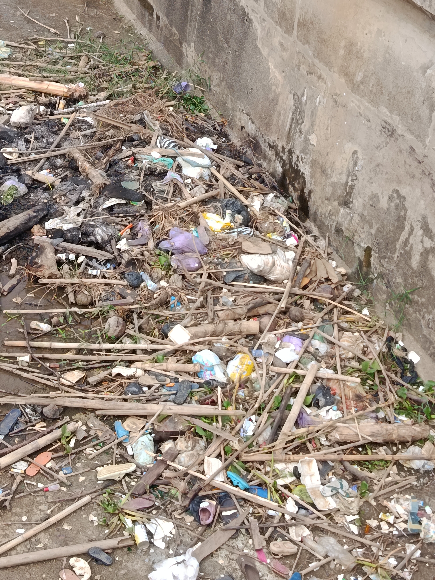 The image shows a large pile of refuse in an outdoor environment, likely a public space or informal settlement in Bantarsari, Indonesia. The refuse consists of a variety of materials including plastic bags, bottles, discarded footwear such as sandals and flip-flops, broken wooden sticks and branches, and organic waste that appears to be partially burned. The ground beneath the refuse is visible in some areas and appears to be dirt or mud. A textured concrete wall, typical of urban infrastructure, forms a boundary on the right side of the image, and some sparse green vegetation is growing amongst the debris and along the base of the wall. There are no people visible in the image. The scene depicts a significant accumulation of unmanaged waste.