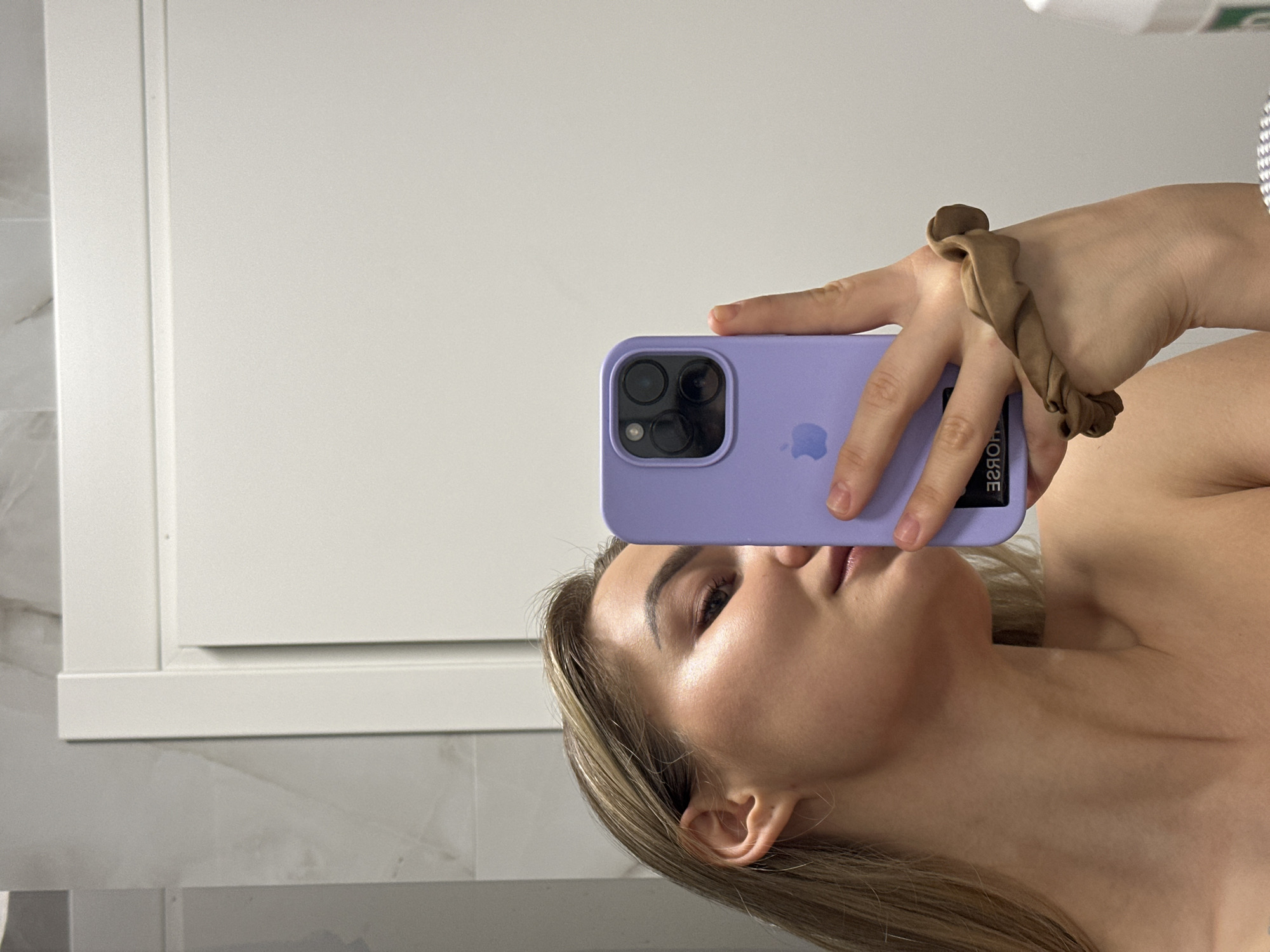 A person is taking a selfie in a bathroom setting. The individual is holding a purple smartphone in front of their face with their left hand. A hair tie is visible on the wrist. The background consists of a white cabinet and light-colored tile. The location is not discernible from the details provided.