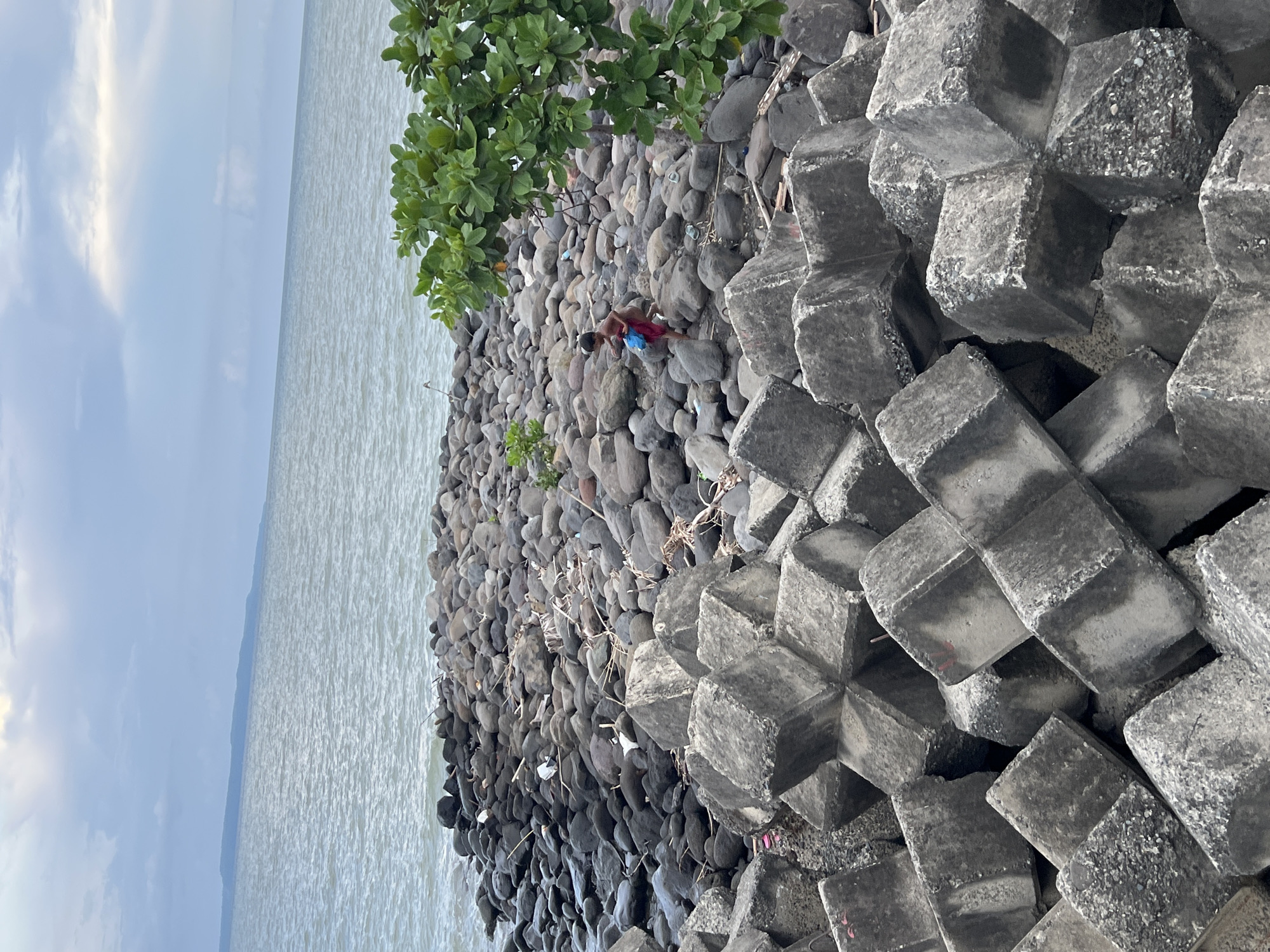 The image depicts a rocky shoreline with a child in the middle ground. The child, wearing red shorts and a blue bag, is walking on the rocks. In the foreground, large concrete tetrapods are stacked, serving as a breakwater. The background shows the sea under a cloudy sky, with a distant landmass visible on the horizon. This scene is likely at a coastal area in Davao City, Philippines, given the presence of similar breakwater structures and the characteristic tropical foliage. The environment appears somewhat unkempt, with debris scattered among the rocks.