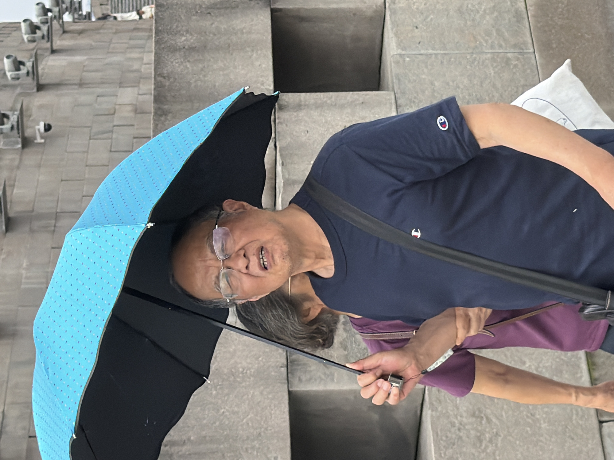 In Yuzhong District, China, an elderly Asian man is holding a bright blue umbrella with a dotted pattern over himself. He is wearing glasses and a dark blue t-shirt with a white logo on the chest and sleeve. His mouth is open, and his facial expression suggests he might be speaking or reacting to something. Partially visible behind him is another person, wearing a purple shirt. They are standing outdoors, with a textured stone wall in the background.