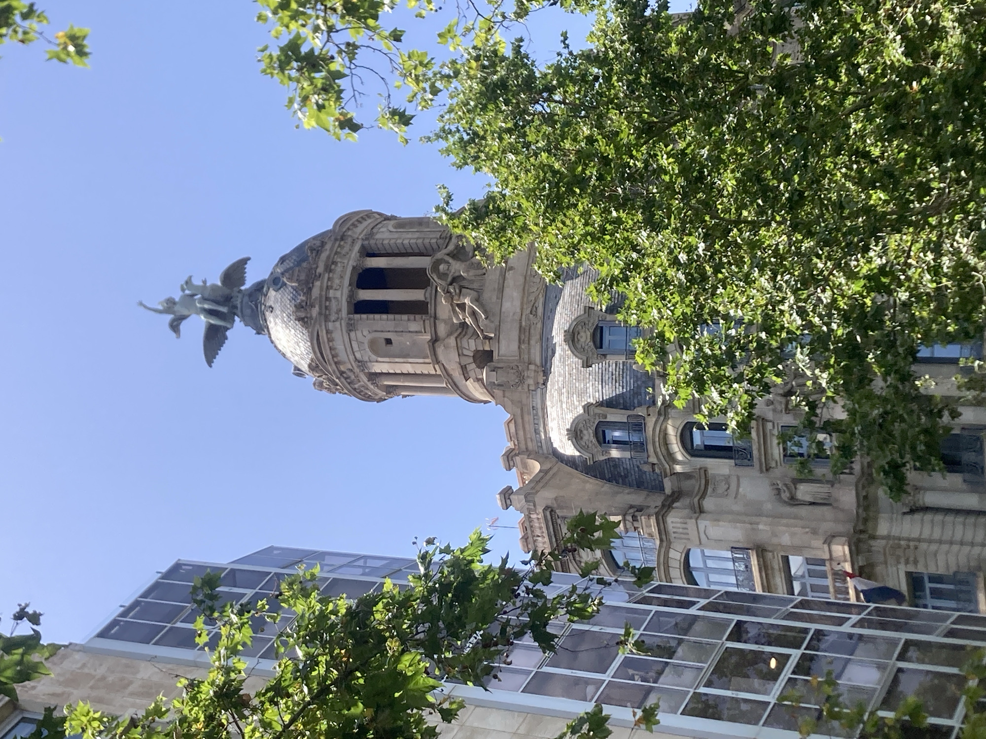 This media file depicts an upward view of an ornate building with a domed tower, partially obscured by lush green trees. The building's architecture suggests historical significance, with intricate stonework and sculptures adorning its facade and the tower. A winged sculpture, possibly an angel, is prominently featured on the upper section of the dome, with its wings outstretched.
Beneath the older building, a modern structure with a glass-paneled roof is visible. The contrast between the classic and contemporary architecture highlights a blend of old and new, likely found in a vibrant urban setting. The clear blue sky suggests it is daytime, and the sunlight filtering through the leaves indicates fair weather.
The scene is set in Barcelona, Spain, a city known for its rich architectural heritage and green spaces. The perspective, looking upwards through the trees, creates a sense of awe and grandeur, inviting the viewer to appreciate the details of the historical building. No people are directly visible, but the presence of the buildings implies an urban environment. The overall mood is one of serene beauty, with the natural elements of the trees complementing the man-made structures. No visible text is present in the image.