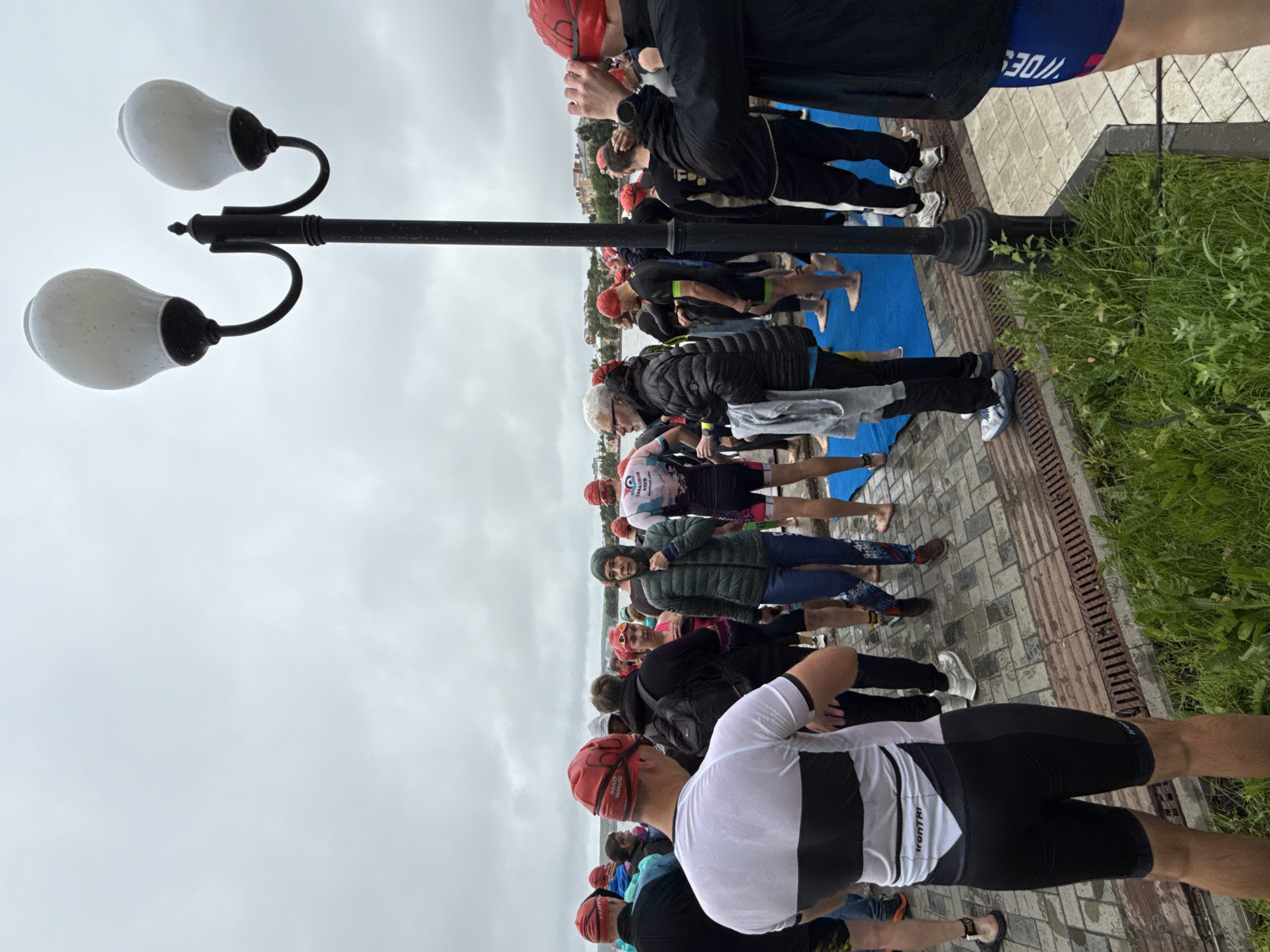 An overcast day in Votkinsk, Russia, with a group of people gathered on a paved walkway near a body of water. Many of the individuals are wearing red swim caps, suggesting they are participants in a water-based event like a triathlon or swimming competition. Some are wearing wetsuits or athletic wear. A black lamppost with two white, frosted glass light fixtures stands prominently in the foreground. To the right, a person wearing a black jacket is adjusting their swim cap, which is red and has "66" written on it. Blue mats are laid out on the ground in front of some of the participants. The environment suggests a public waterfront area, possibly a park or embankment. In the background, buildings and trees are visible across the water. The ground is paved with light-colored stones, and there are metal grates along the edge of the walkway. Lush green grass grows along the edge of the paved area.
