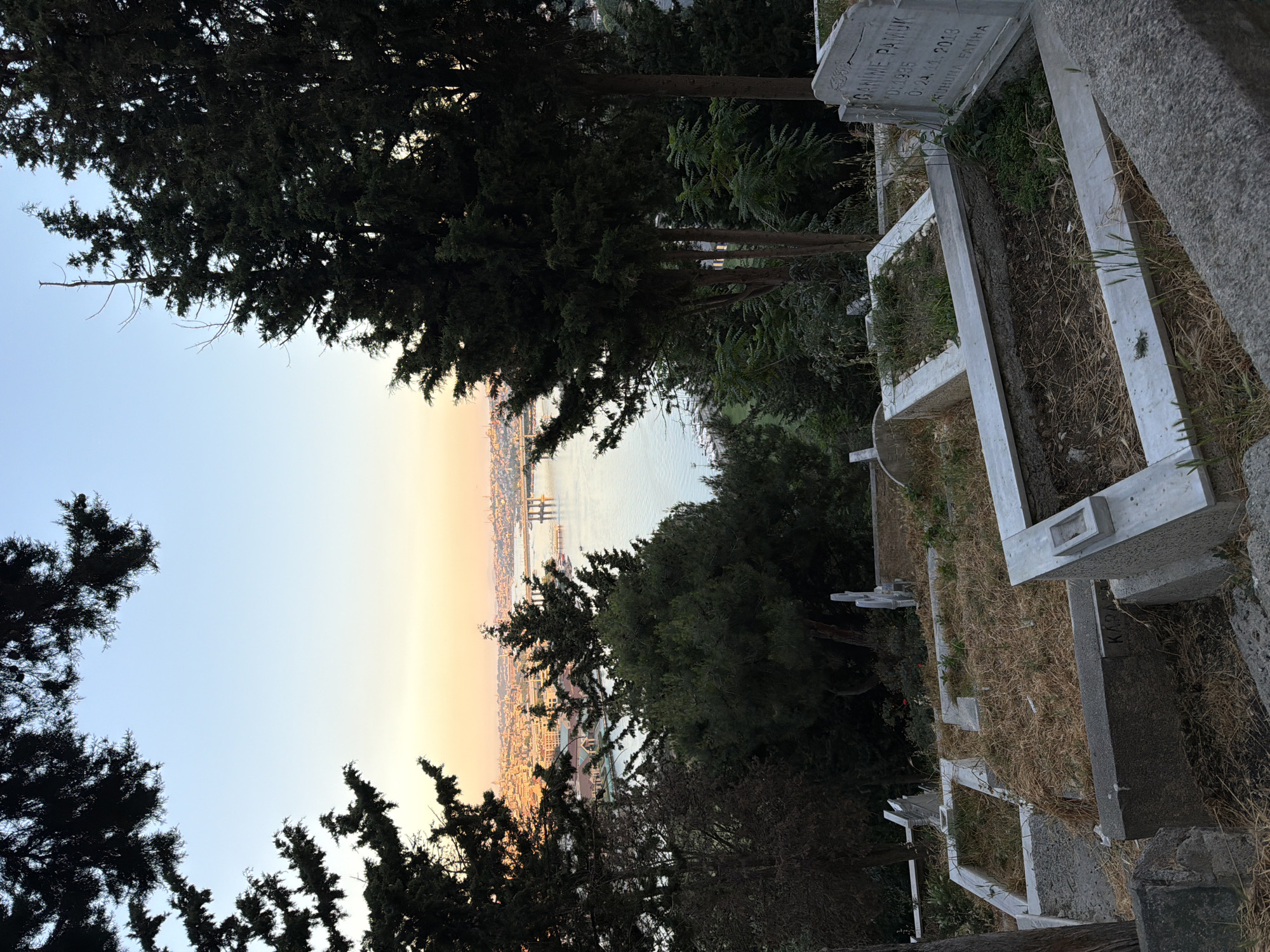 The image depicts a cemetery in Eyüpsultan, Turkey, overlooking a body of water and a city skyline at dusk. In the foreground, several gravestones, some with white marble detailing, are visible amongst dry grass and sparse vegetation. A prominent gravestone on the right side bears an inscription in Turkish, with the name "GANIME PAMUK" and dates "DE: 1935" and "O: 24. 11. 2013" legible. The middle ground is a densely wooded area with tall evergreen trees. Beyond the trees, a wide body of water, likely the Golden Horn, stretches towards the horizon. The far background reveals a sprawling city with buildings bathed in the warm hues of sunset. A bridge and various structures are discernible along the waterfront. The sky above is a gradient of pale blue to soft orange and yellow, indicating the end of the day. There are no people or animals present in the scene.