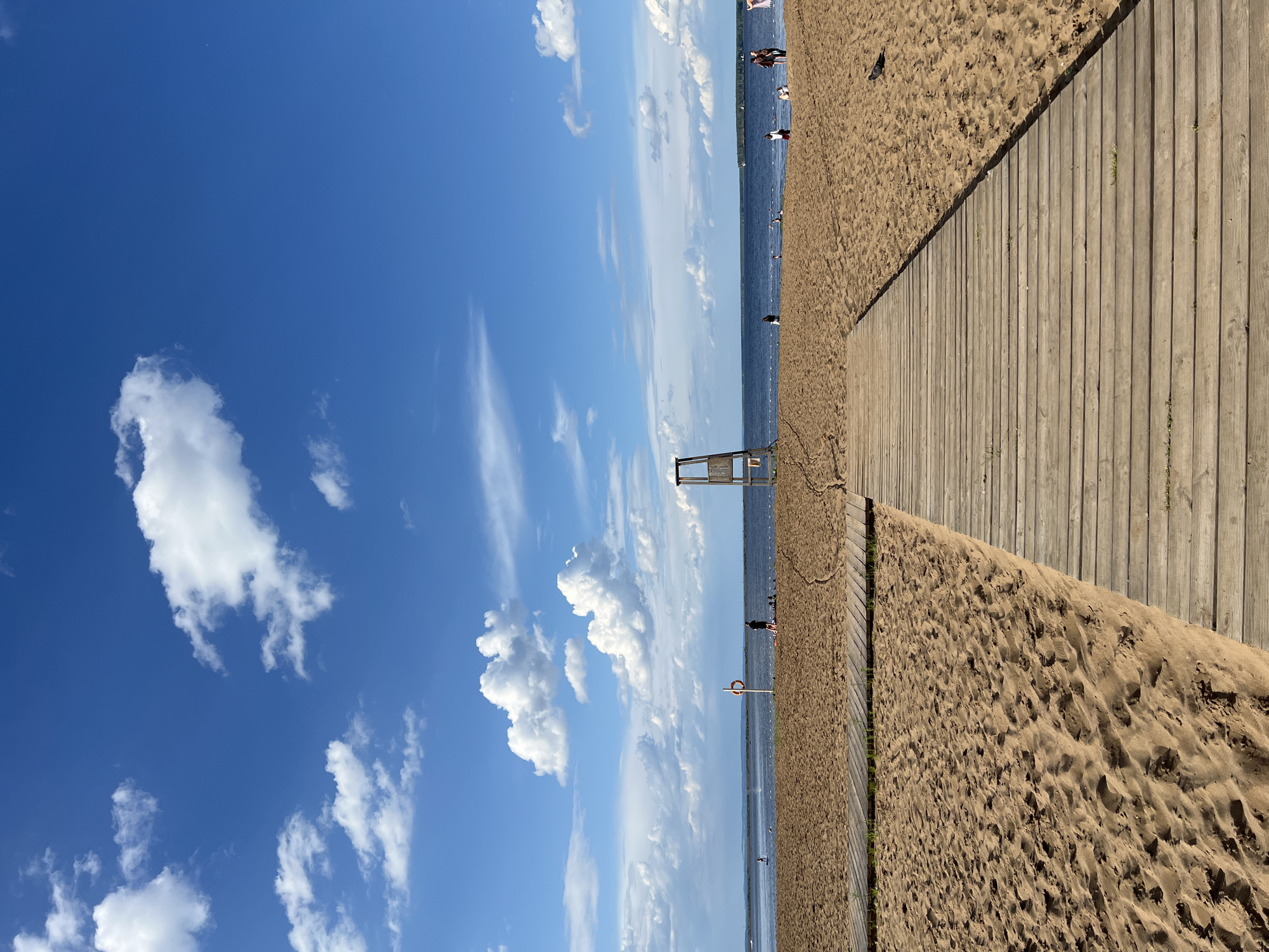 A wooden boardwalk leads across a sandy beach towards a calm blue body of water. White, fluffy clouds dot the vibrant blue sky. On the beach, several people are visible, some walking and others lounging. A lifeguard tower stands prominently near the water's edge, and a red life preserver hangs on a pole. The scene captures a serene summer day at the beach.