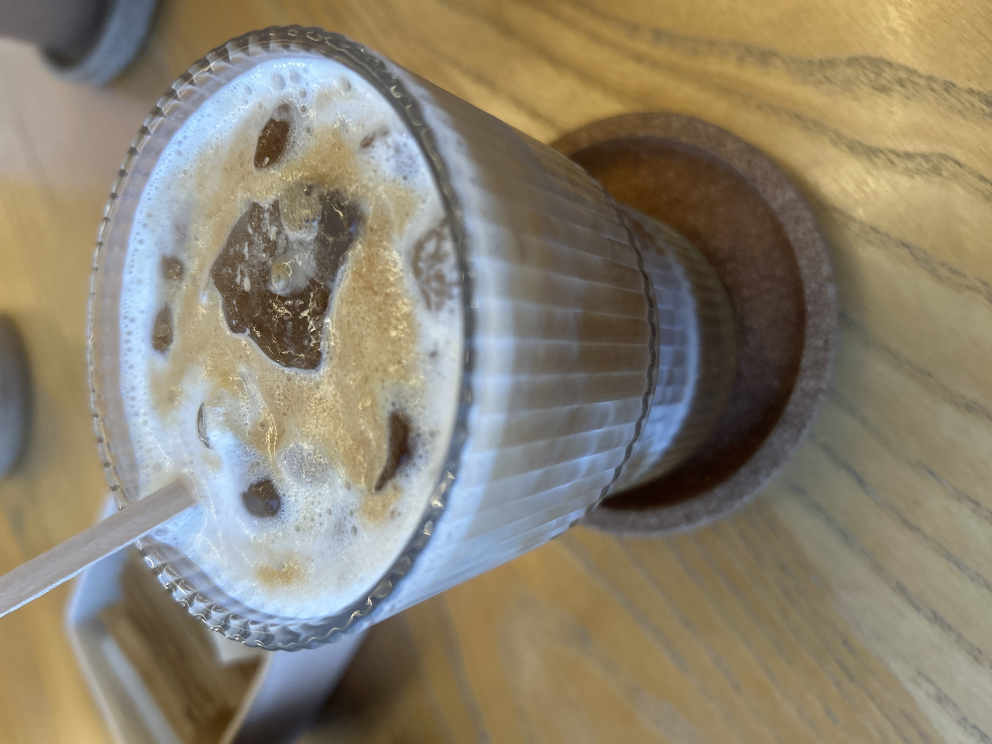 An overhead, slightly angled shot captures a refreshing iced beverage served in a clear, vertically ribbed glass. The drink appears to be an iced coffee or a similar layered cold beverage, showcasing distinct strata: a lighter, milky base beneath a darker coffee layer, topped with a frothy, bubbly cream or foam. Several irregularly shaped ice cubes are interspersed within the liquid and froth. A light-colored, possibly paper or wooden, straw extends into the drink from the lower left side of the frame, indicating readiness for consumption.
The glass rests on a round, textured coaster, likely made of cork, which provides a contrasting earthy tone against the lighter elements of the drink. Both the glass and coaster are positioned on a wooden table, characterized by its warm, light brown hue and pronounced, dark wood grain patterns that stretch horizontally across the surface. The lighting is soft and even, highlighting the textures of the drink's foam and the glass, and casting gentle shadows. The overall setting suggests a calm indoor environment, possibly a cafe or home, focused on the enjoyment of the beverage. There is no visible text in the image.