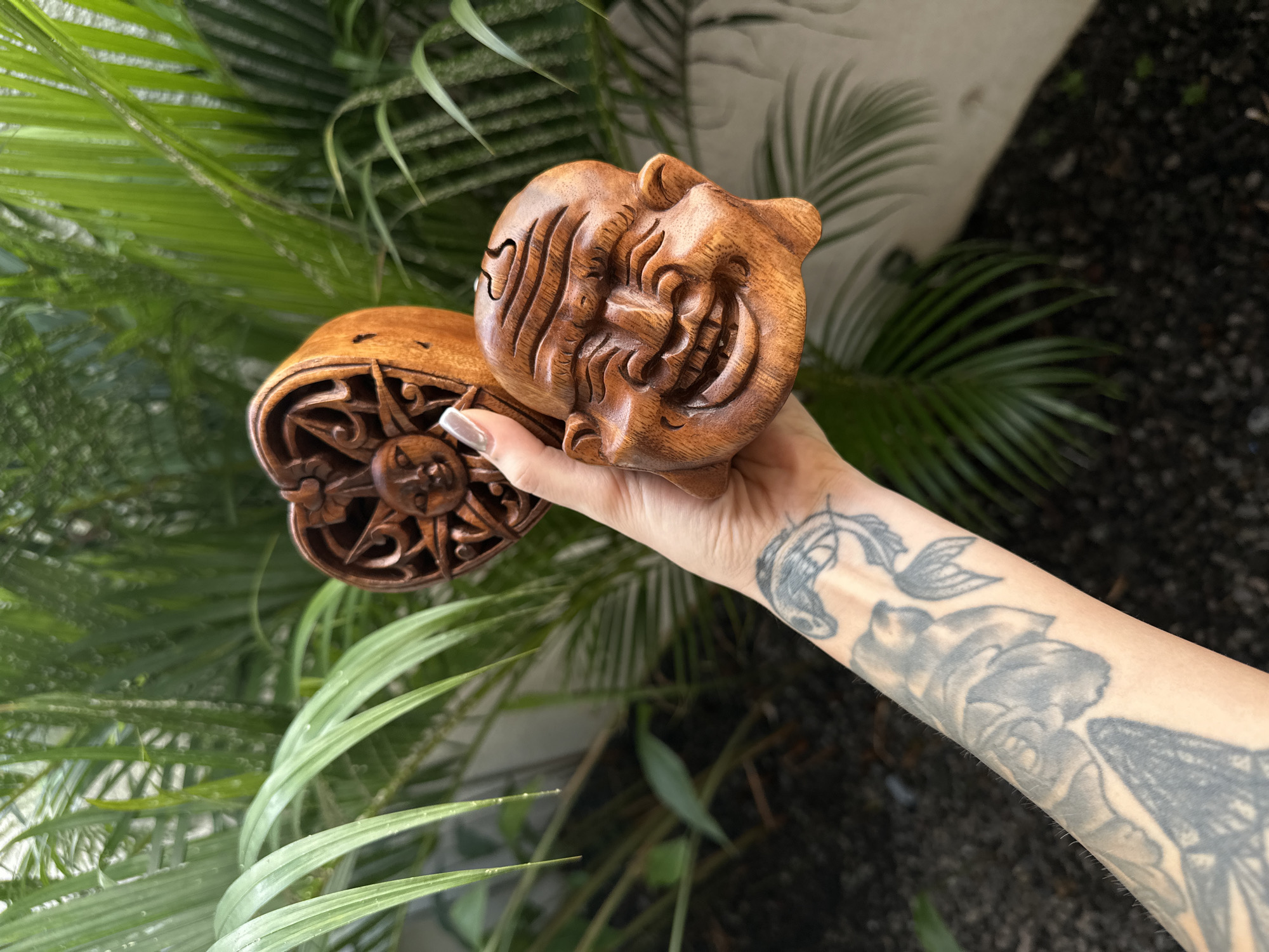 A close-up, outdoor shot captures a person's tattooed arm and hand holding two intricately carved wooden objects against a vibrant green tropical background, typical of North Kuta, Indonesia.
The primary object is a rich brown wooden carving depicting a highly expressive, almost caricatured face with wide, elongated eyes and a broad, toothy grin. Its features are deeply carved, suggesting a traditional or mythical figure, possibly part of a puzzle box or container. The second wooden piece, held underneath the first, is circular or heart-shaped and features a detailed sun-like motif surrounded by ornate, swirling patterns.
The hand holding these items has well-manicured nails with a light, shimmery polish. The forearm is extensively tattooed with several designs, including what appears to be a stylized fish or mermaid tail near the wrist, a large, intricate floral pattern (possibly a rose), and other abstract or organic shapes in shades of grey and black.
The background is dominated by bright green palm leaves and other tropical plants, indicating an outdoor, sunny environment. A light-colored wall and dark ground cover are visible behind the foliage. The scene appears to be taken during the daytime, showcasing the detailed craftsmanship of the wooden carvings and the personal artistry on the arm. There is no visible text in the image.