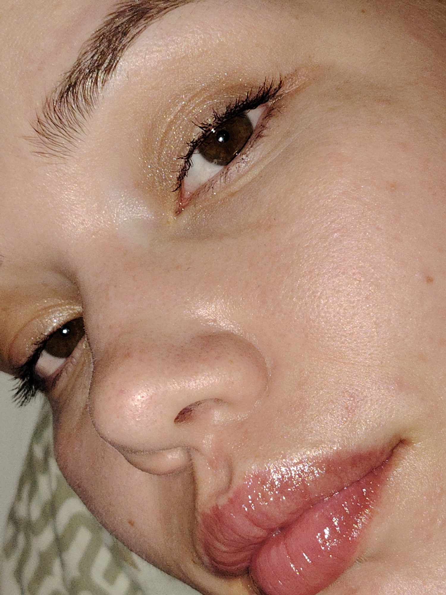A close-up, angled photograph, taken in Марковский, Russia, depicts the right side of an individual's face. The subject exhibits fair skin with visible pores and minor blemishes. The right eye is open, revealing a dark brown iris and a dark pupil. The upper eyelid features a light-colored, shimmering eyeshadow with glitter particles. Eyelashes, coated with mascara, are present on both upper and lower lids. The right eyebrow is well-defined with brown hairs. The bridge and tip of the nose are partially visible, angled downwards and to the left. The lips, positioned in the lower right, are full, glossy, and coated with a reddish-pink cosmetic product, appearing slightly parted. The overall expression is neutral. The background, out of focus, reveals a light-colored fabric with an indistinct patterned texture, suggesting an indoor setting.