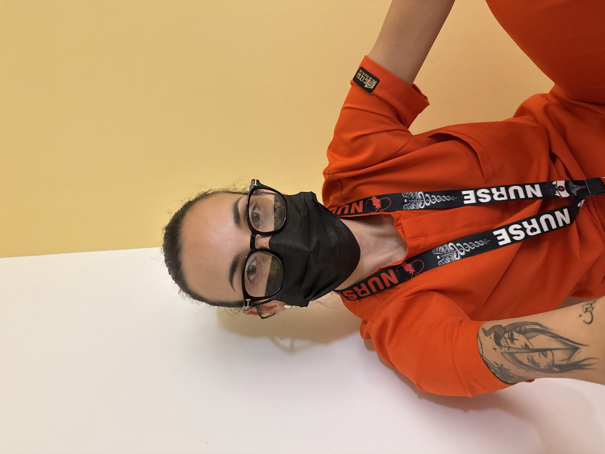 An individual, likely a woman, is captured in a close-up, tilted portrait. She wears an orange scrub top and a black face mask, with black-framed glasses resting on her nose. Her dark hair is pulled back, and she looks directly at the camera with a neutral expression. A black lanyard featuring white "NURSE" text interspersed with a medical caduceus symbol hangs around her neck. On her right arm, a prominent black-and-white tattoo depicts a stylized face or figure, and a smaller tattoo resembling an Om symbol is visible on her upper arm. The cuff of her orange sleeve has a label that reads "CİZGİ". The background is a simple two-toned wall, with a mustard yellow hue on the upper half and white on the lower half, divided by a distinct horizontal line. The lighting is bright and even, suggesting an indoor setting.