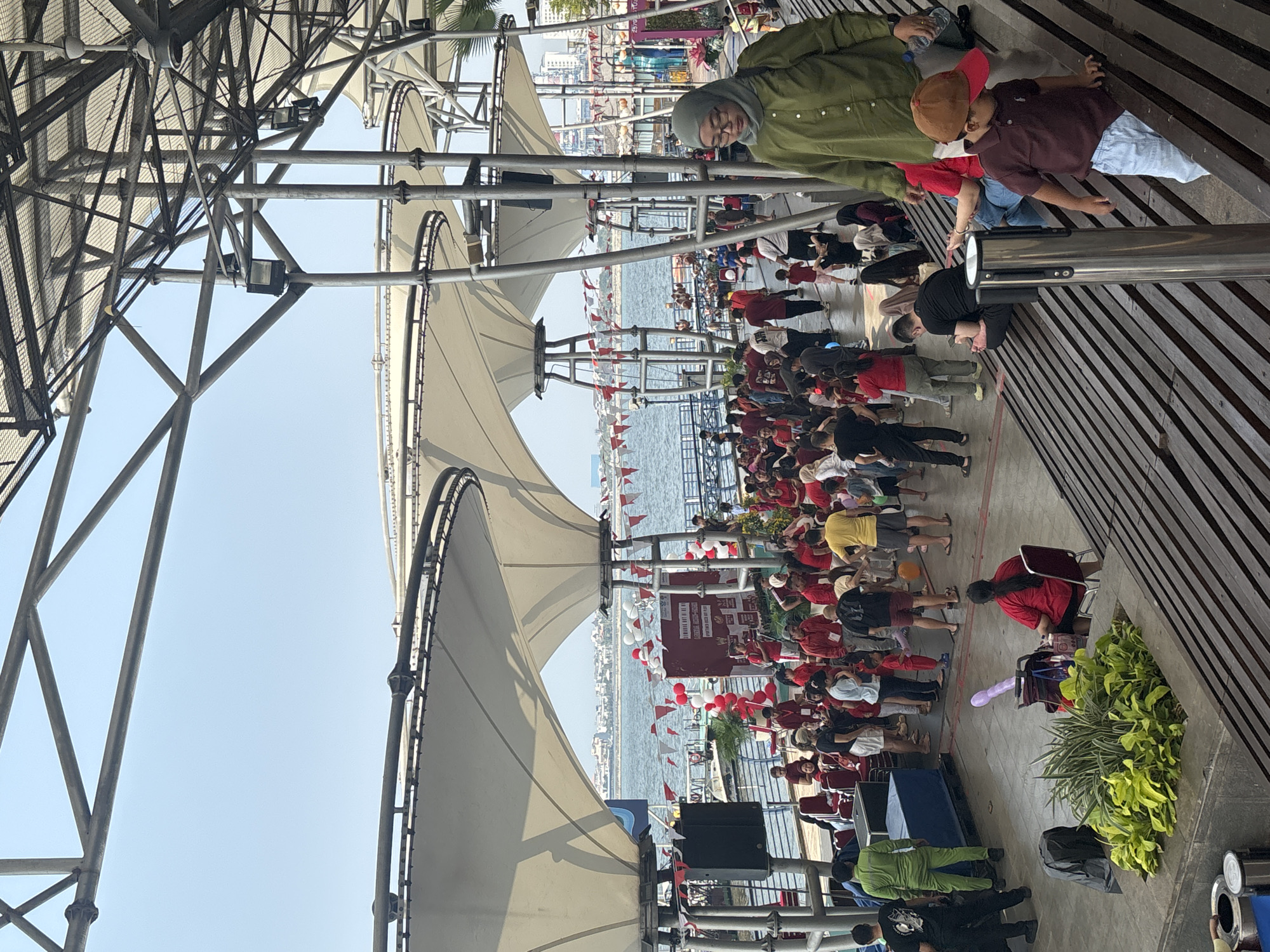 The image depicts a crowded outdoor event at Ancol Beach City in North Jakarta, Indonesia. A large, white canopy structure covers part of the scene, with metal framework visible above. In the background, the sea is visible, with a distant cityscape on the horizon.
A diverse crowd of people, mostly dressed in red and white, are gathered. Many are standing, while some are seated on tiered wooden benches. There's a sense of activity and festivity, with balloons and flags decorating the area. Some individuals appear to be interacting with each other, while others are observing the surroundings.
Objects in the foreground include a large speaker system, red chairs, a blue storage bin, and a potted plant. A child wearing a brown cap and red collar shirt is walking down the wooden steps. A woman wearing a green headscarf and glasses is also visible on the steps. The overall environment suggests a public gathering or festival.