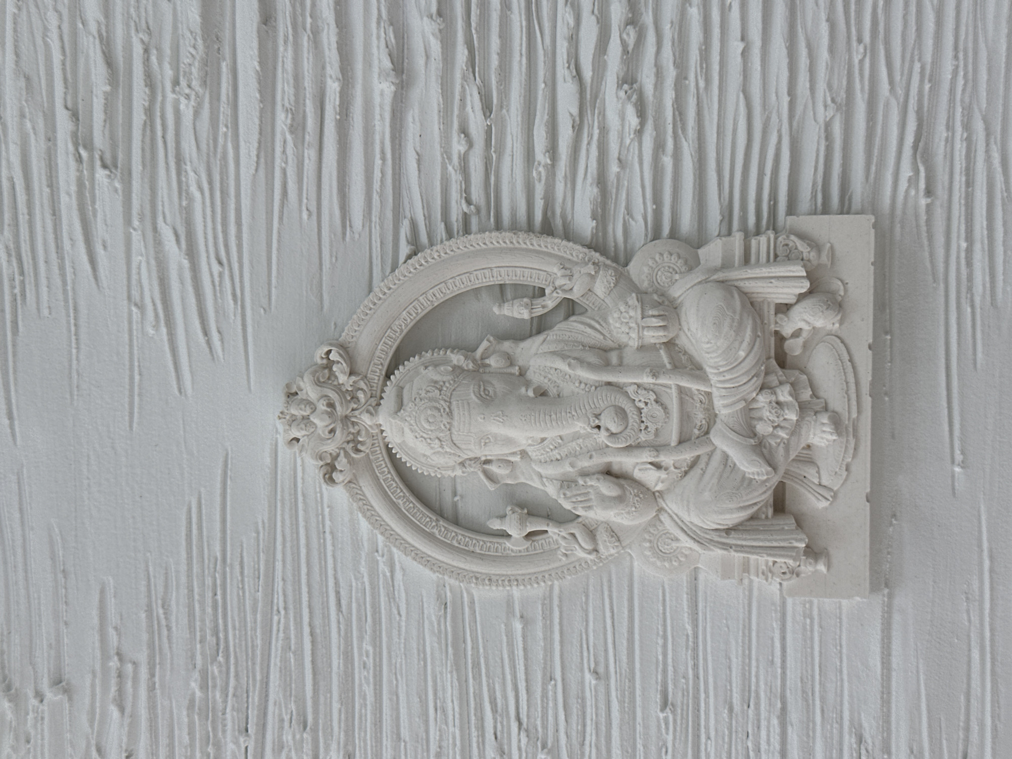 The image shows a white bas-relief sculpture of the Hindu god Ganesha, mounted on a textured white wall. The sculpture is detailed, depicting Ganesha in a seated pose within an ornate oval frame. The texture of the wall behind the sculpture has vertical lines. The location is not provided, and there is no definitive evidence to suggest this image was taken in Sochi, Russia.