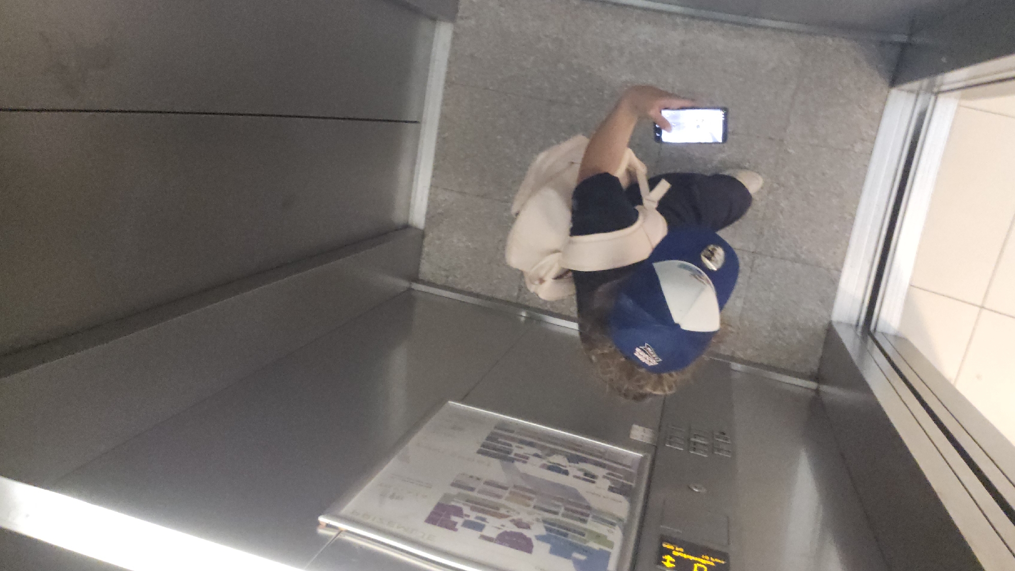 A high-angle shot looking down inside an elevator. A person, identifiable by their partially visible clothing (navy blue jacket, white bag), is standing in the elevator, holding a smartphone. The elevator's interior is metallic, with a control panel and a floor map visible on the lower part of the frame. The elevator door is open, revealing a brightly lit interior space beyond. The location of Unknown, Unknown, is not determinable from the image.