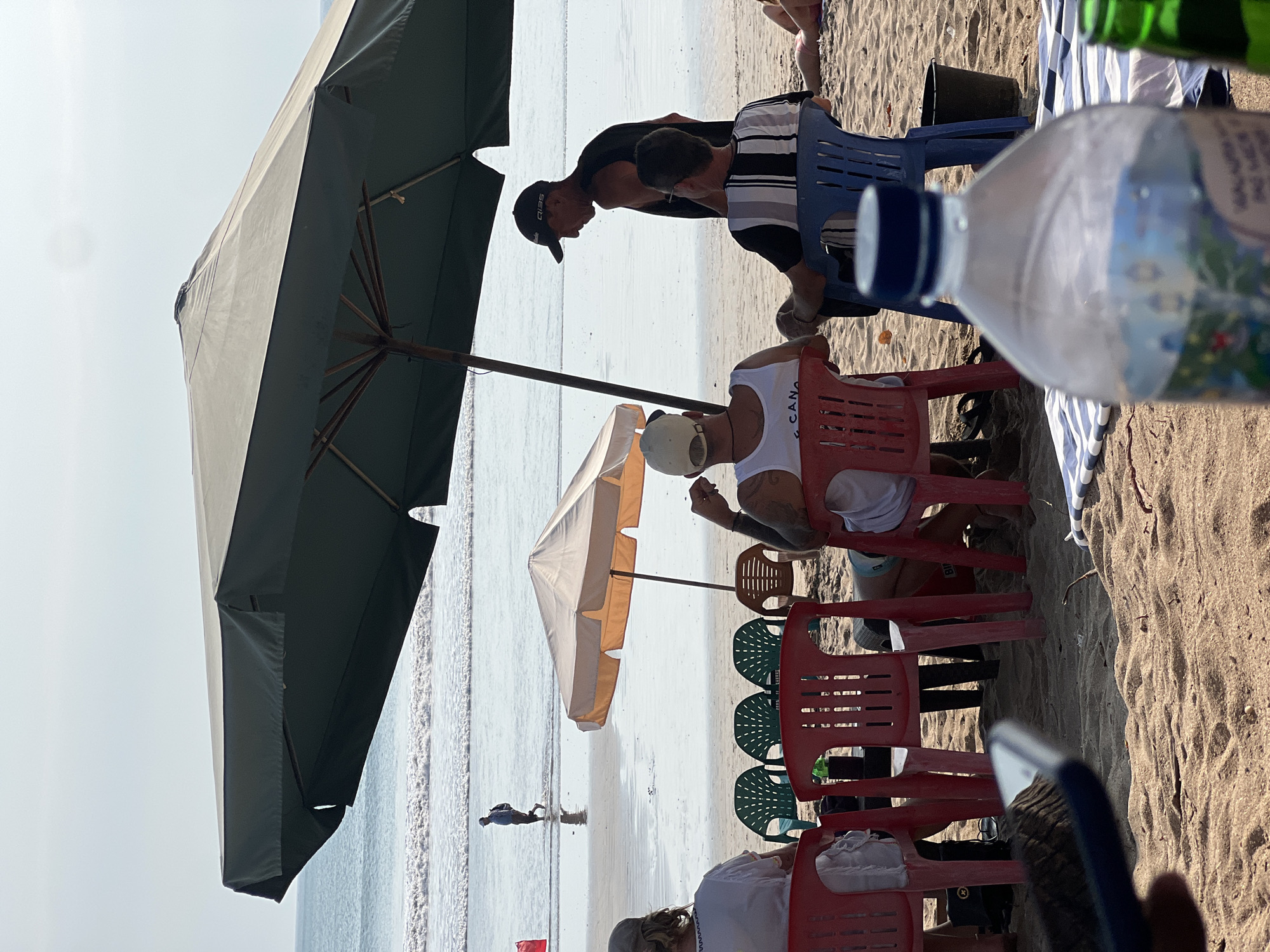 The image shows a group of people sitting on a beach in Kuta, Indonesia, under a large dark umbrella. The beach is sandy, with the ocean and waves visible in the background. The people are seated on plastic chairs, and there are other beach umbrellas and chairs around. One person is walking in the water near the shoreline. The scene suggests a casual gathering on a sunny day at the beach.
