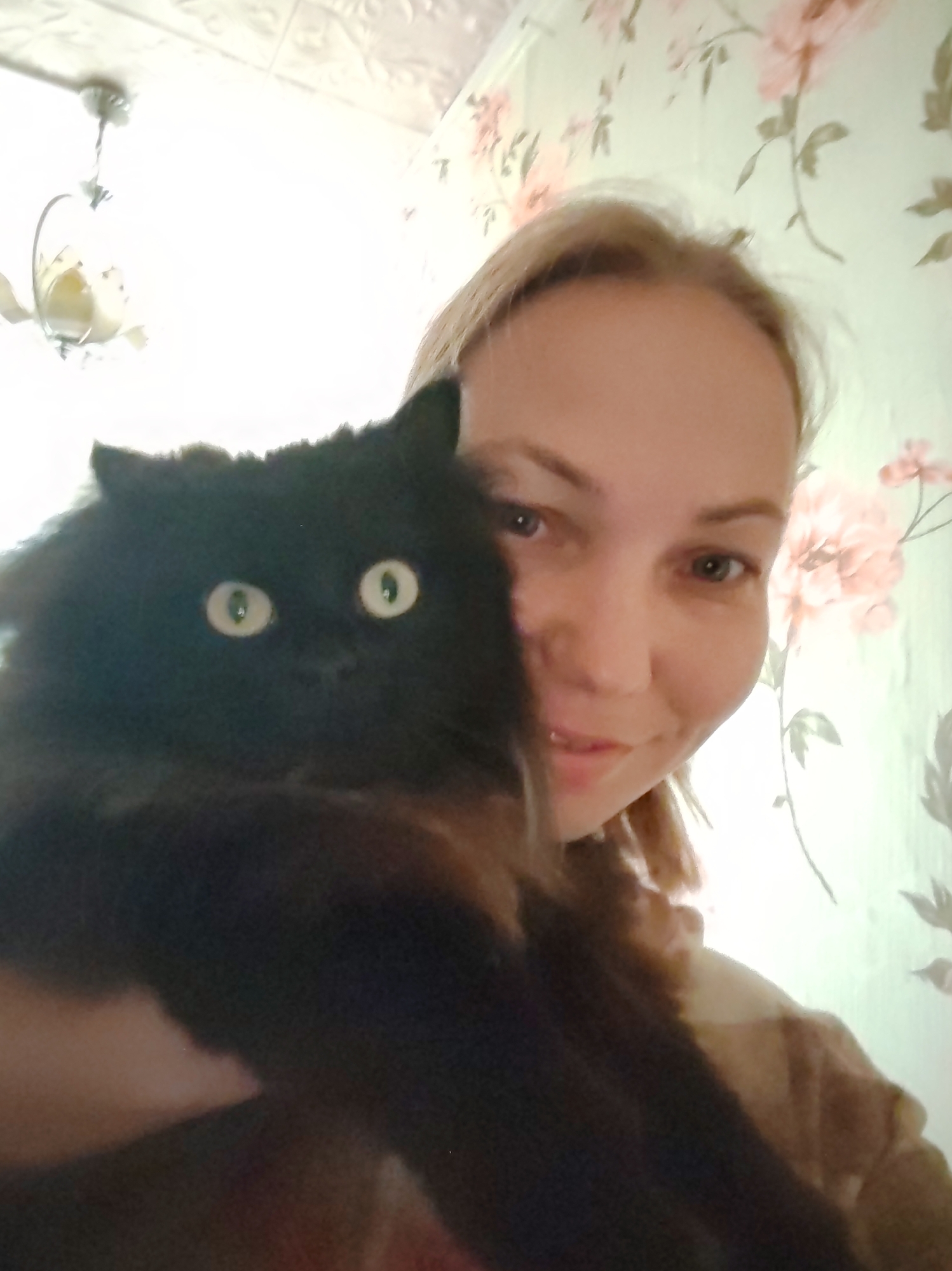 A woman with blonde hair is holding a black cat in front of her. The cat's eyes are wide and staring directly at the camera. The woman is smiling faintly. They are in a room with floral wallpaper and a chandelier visible in the background. The scene appears to be a selfie taken indoors, possibly in a home. The woman is wearing a light-colored top. The cat has long, fluffy fur. The lighting is bright, and the image has a slightly blurry quality. The overall impression is one of casual intimacy between the woman and her pet.