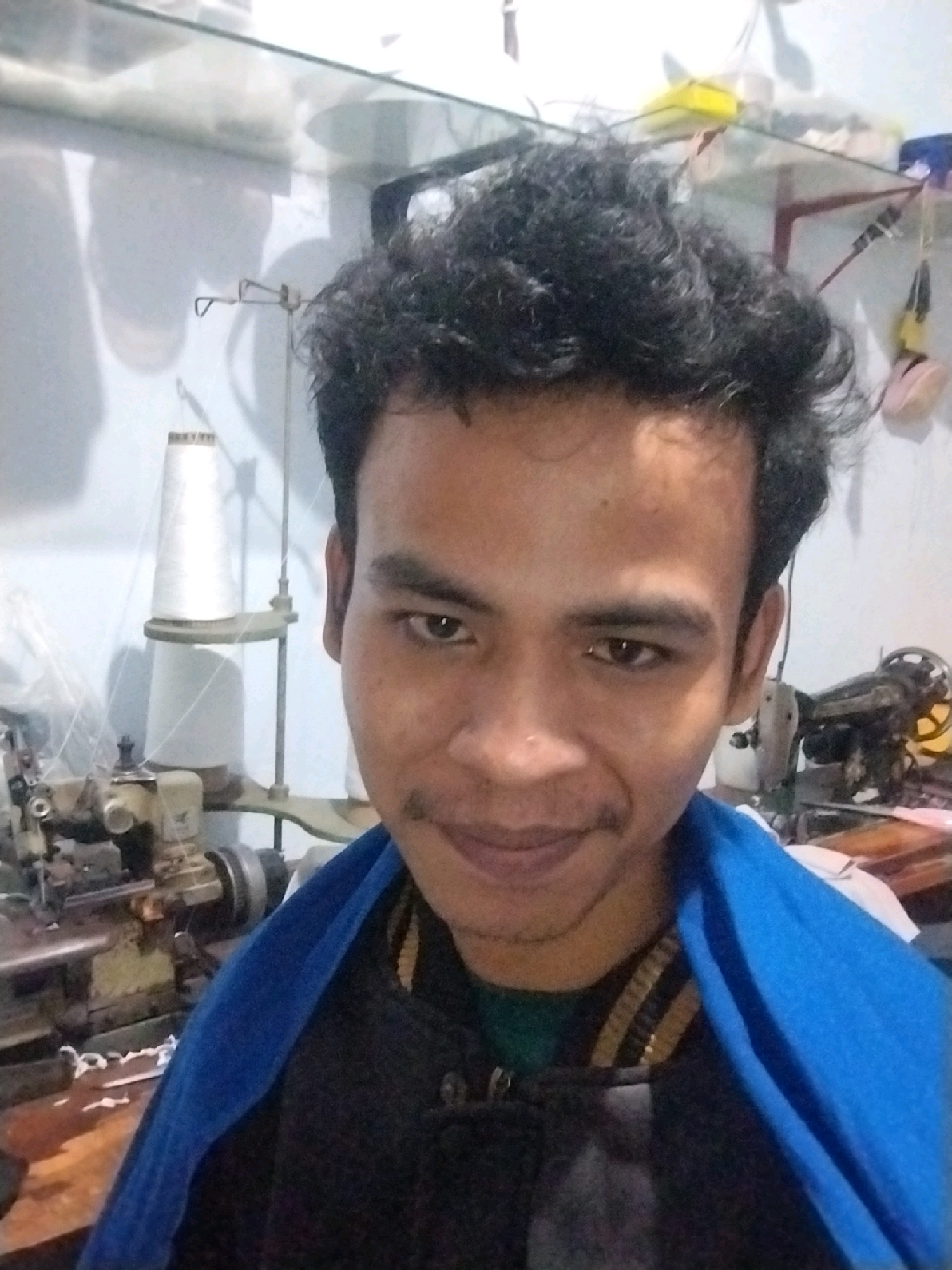 A young man with curly black hair and a faint mustache smiles slightly at the camera. He is wearing a blue garment draped over his shoulders, with a black and yellow striped shirt visible underneath. He appears to be in an indoor workshop. To his left, a sewing machine is visible, along with a large white spool of thread on a stand. The background shows shelves with various items and a second sewing machine to the right. The lighting is dim, suggesting artificial light sources. The overall environment suggests a sewing or tailoring business, possibly located in Mumbulsari, Indonesia, given the context.