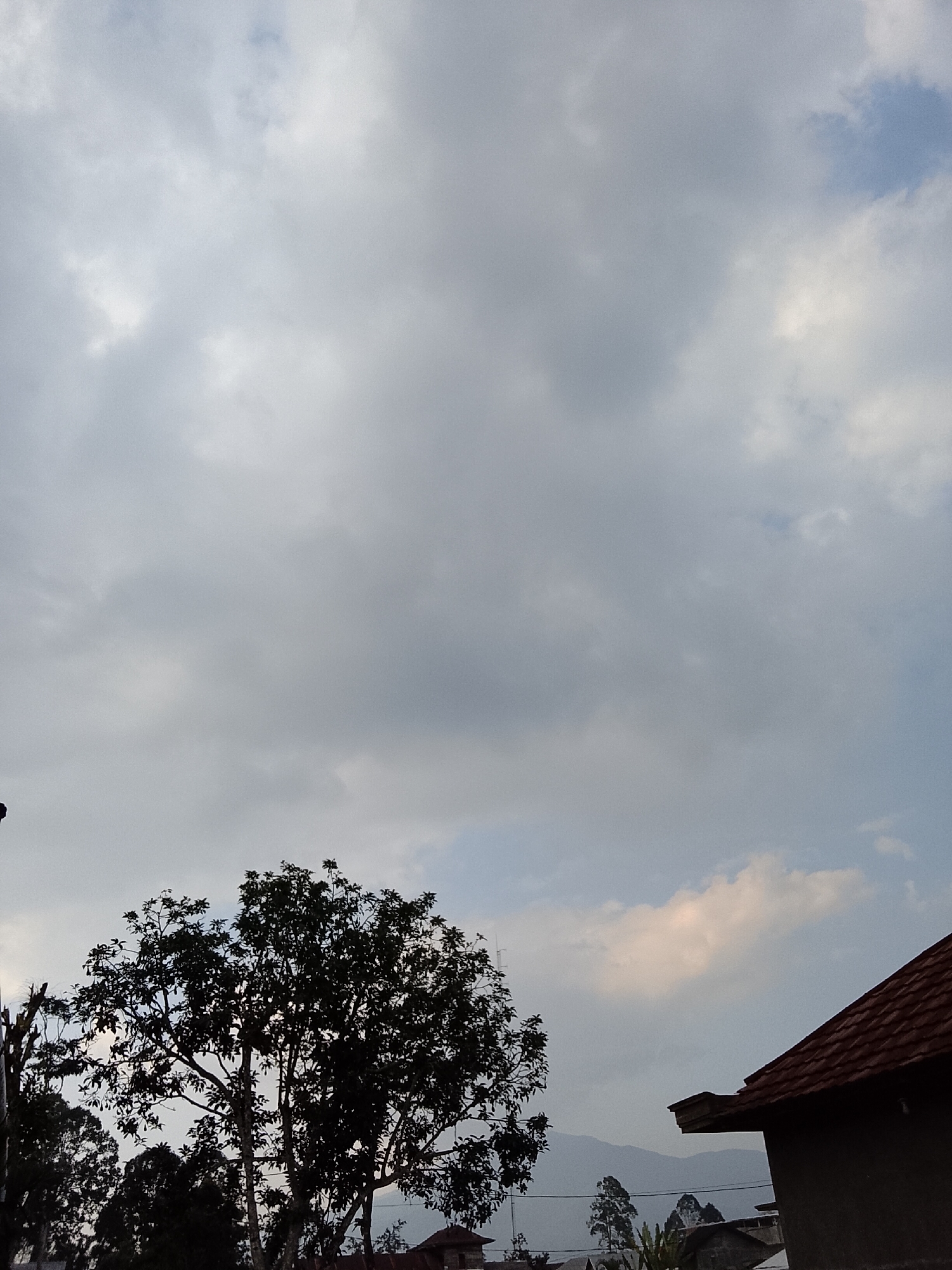 The image depicts a cloudy sky over a landscape that appears to be in Padang Dalom, Indonesia. In the foreground, the silhouette of a large tree with dense foliage is visible on the left. To the right, a portion of a building with a brown, tiled roof is present. In the background, distant mountains are faintly visible through the hazy sky, along with other trees and structures. The overall impression is a serene, natural setting under an overcast sky.