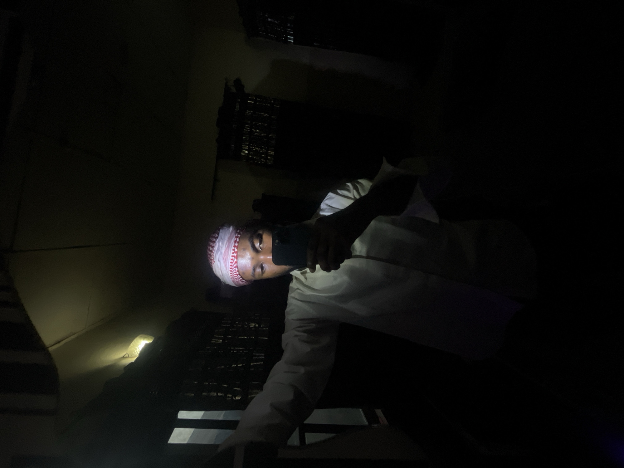 A person wearing a white shirt and a red and white patterned head covering is holding a mobile phone in a dark environment. The location appears to be a room with window coverings, possibly in a city with high-rise buildings visible through the window.