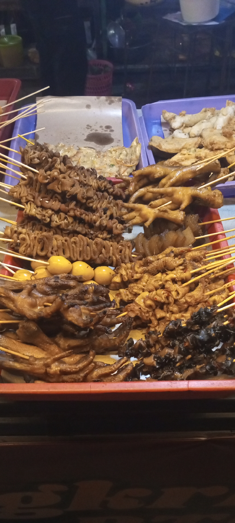 In Setu, Indonesia, a street food stall displays an assortment of skewered and fried animal parts for sale. The food is arranged in red and purple plastic trays. Visible items include intestines, chicken feet, quail eggs, and what appears to be fried chicken. The food is lit by overhead lights, creating a slightly blurry and high-contrast image. In the background, other market stalls and possibly shoppers can be seen. The overall impression is a vibrant, bustling night market scene.