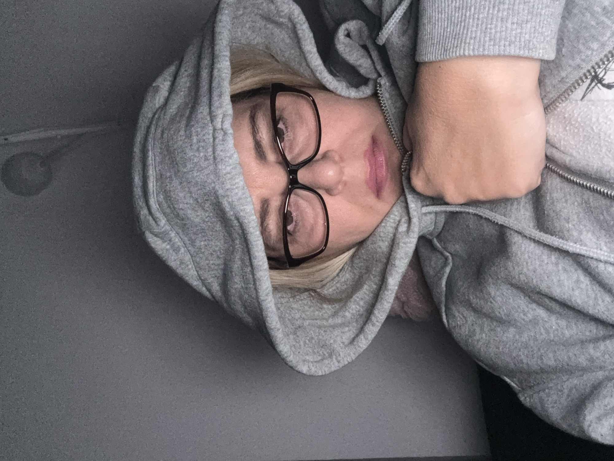 A person wearing a gray hooded sweatshirt and black eyeglasses is shown. The person's face is partially obscured by the hood. The background appears to be a gray or dark-colored wall, possibly indoors. A hand is near the zipper of the sweatshirt. A light is on the left wall.