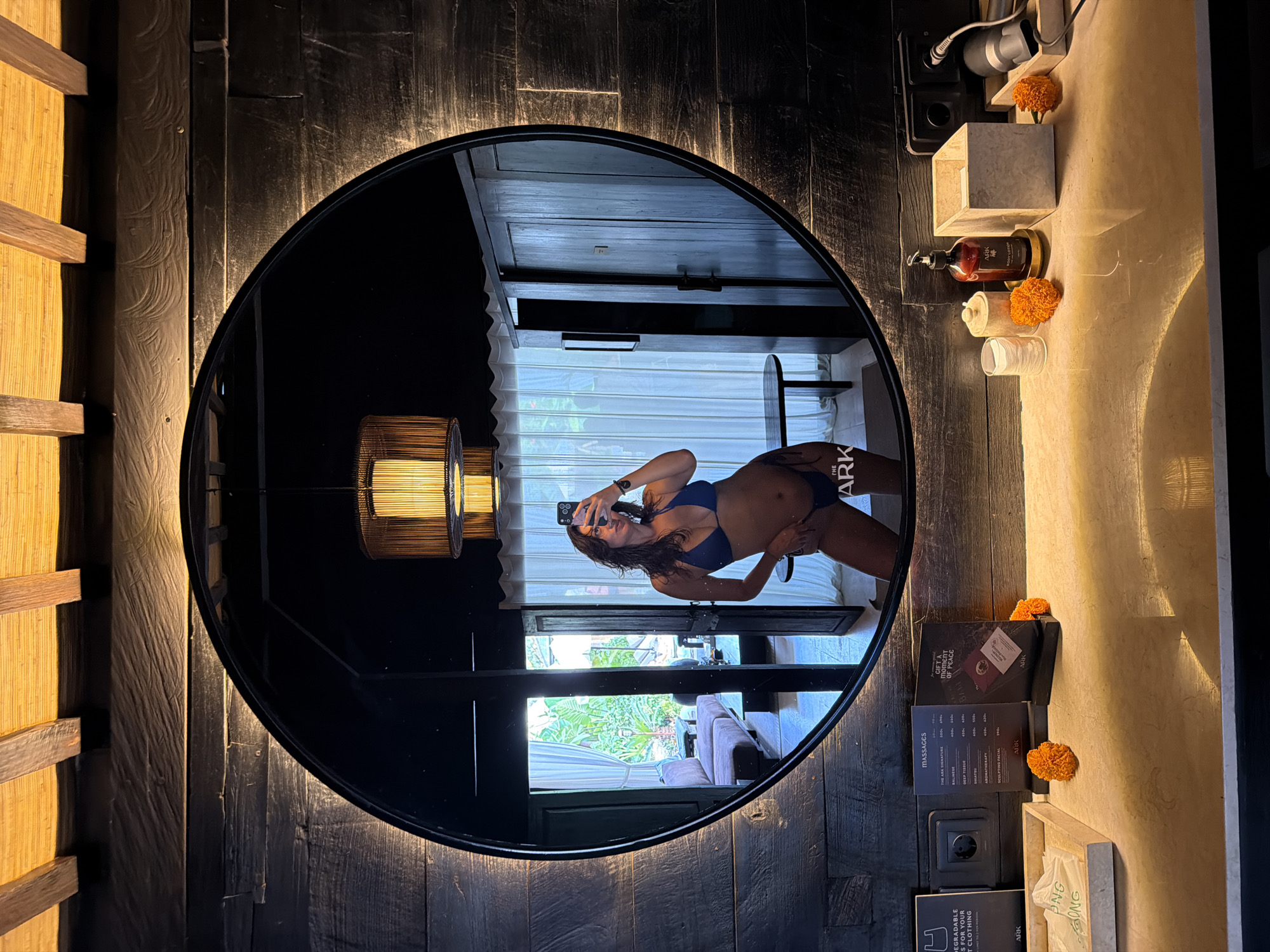 A woman, viewed in a mirror's reflection, stands in a bathroom. She is wearing a blue bikini and holding a smartphone to take a selfie. Her reflection shows her from the mid-thigh up. The bathroom has a rustic aesthetic with dark wood paneling and exposed wooden ceiling beams. A large, circular mirror is mounted on the wall. On the counter below the mirror are various items, including toiletries, decorative flowers, and a menu. The room behind her, visible in the mirror, appears to be a bedroom with a sofa, table, and open doors leading to an outdoor area. The image was taken in Mengwi, Indonesia, at a resort.