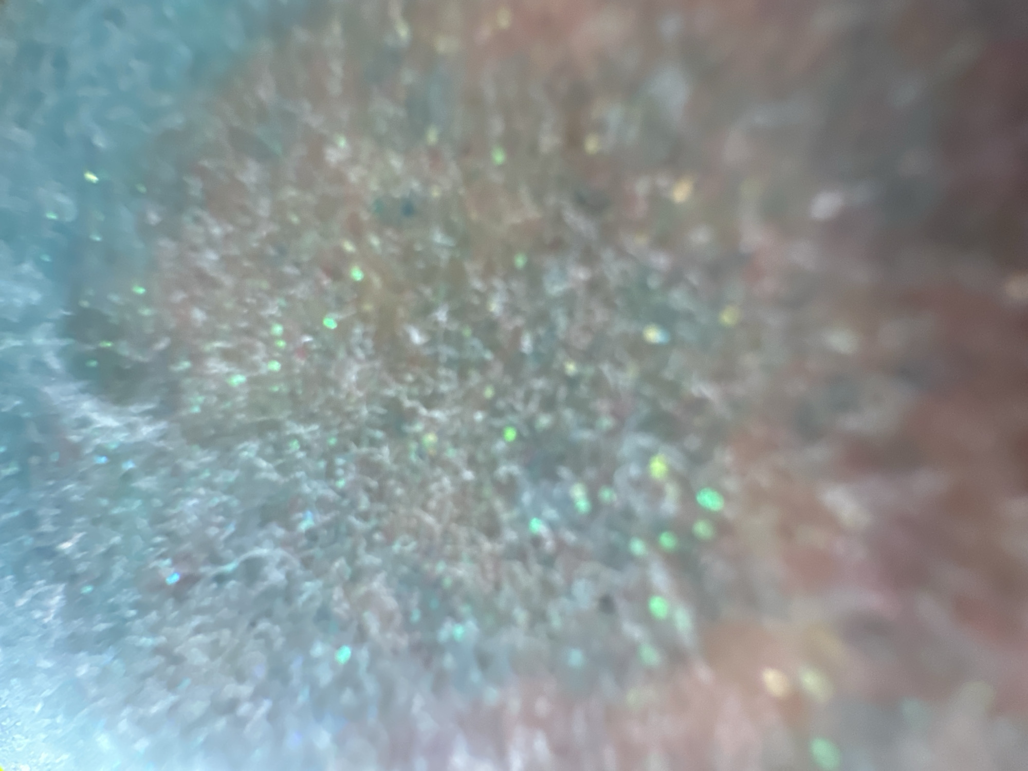 This is a close-up, out-of-focus abstract image featuring a glittery texture. The colors are predominantly light blue, silver, and soft pinkish-brown, with small specks of green and yellow light reflecting throughout. The overall effect is shimmering and iridescent, suggesting the presence of small particles that catch and refract light. There are no discernible people, objects, or a clear scene or setting. The activity or event taking place is not evident due to the abstract nature of the image. No time of day, weather, location cues, or emotions can be determined. There is no visible text.