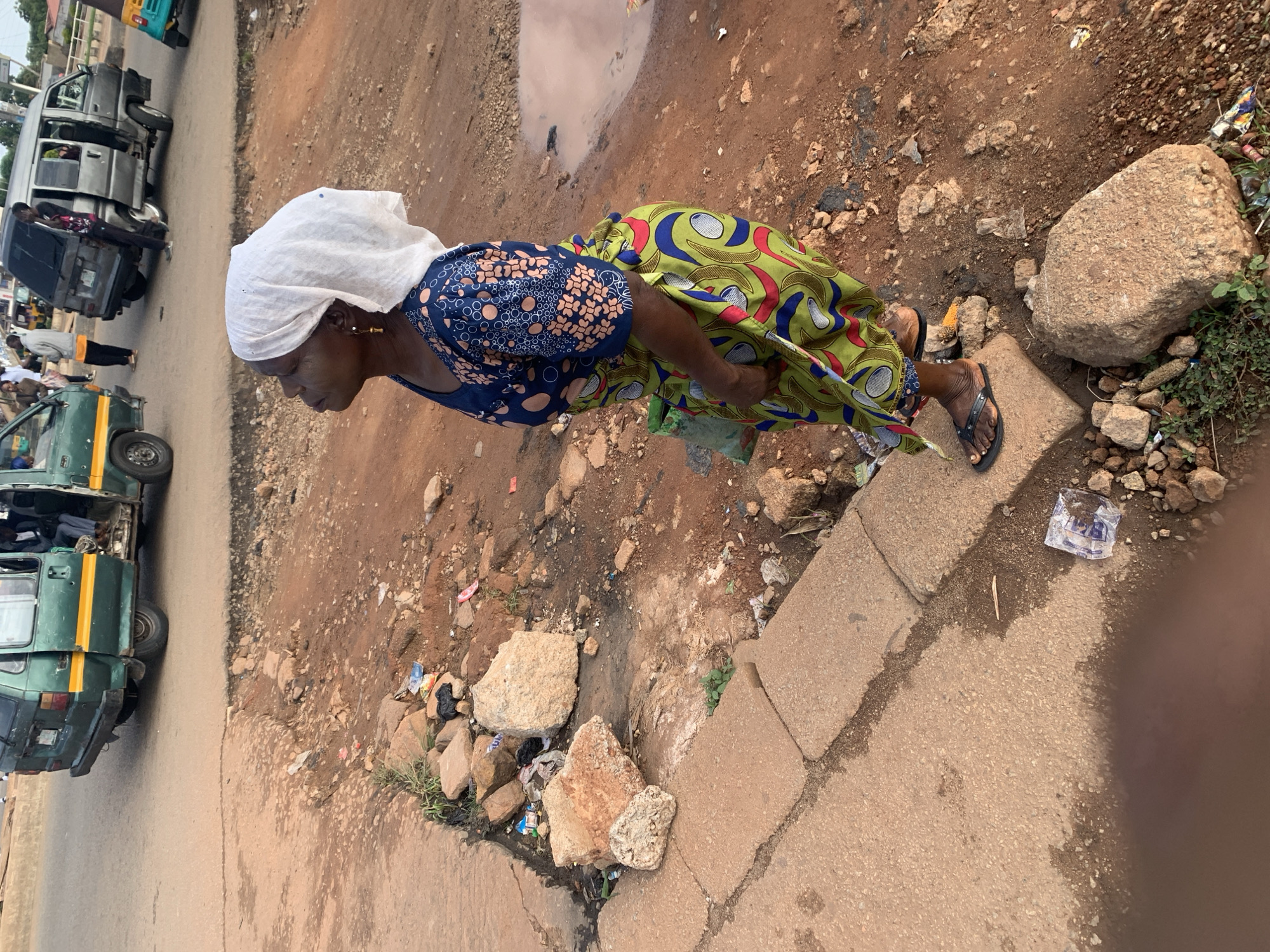 In Gura Top, Nigeria, a woman stands on a dusty dirt road next to a patched sidewalk. She wears a white headscarf and a patterned blue and orange dress, with a colorful green and red patterned wrap skirt. Her feet are clad in black flip-flops. Behind her, a weathered green minivan with yellow trim is parked, its door open, revealing passengers inside. Further back, a silver minivan and a turquoise tricycle are visible on the road. The environment is characterized by dry, reddish-brown earth, scattered rocks, and some debris, including discarded plastic bags and wrappers. A small puddle of muddy water is present on the ground. The scene appears to be a street-side waiting area or a roadside stop.