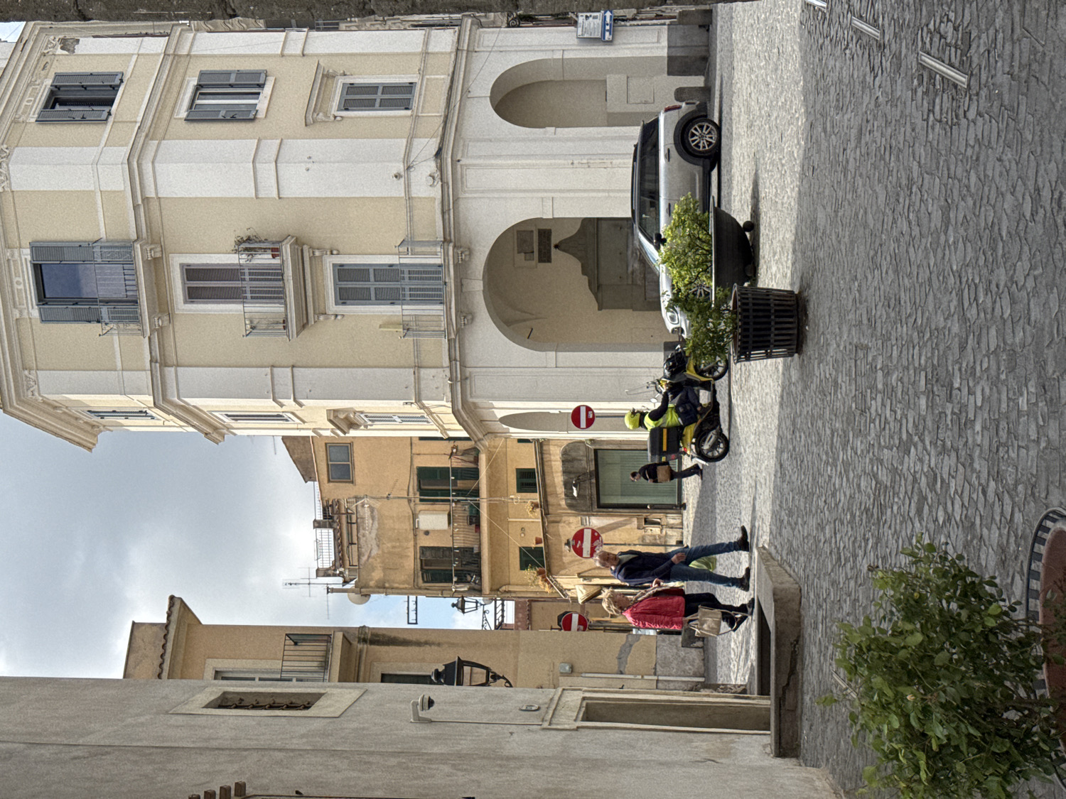 The image captures a daytime street scene in Ariccia, Italy, under an overcast yet bright sky. The street is paved with distinctive grey cobblestones and flanked by multi-story buildings of varying architectural styles.
To the left, a prominent building features a facade of alternating cream and white horizontal bands, adorned with numerous windows, many fitted with closed grey louvered shutters. Its ground floor is characterized by large arched passageways, beneath one of which a silver SUV is parked.
In the mid-ground, several individuals populate the street. A couple walks towards the viewer; the man wears a dark blue jacket and jeans, while the woman is in a bright red jacket, carrying a light-colored handbag. Further down the street, a person in a high-visibility yellow jacket and helmet is seated on a bright yellow scooter, suggesting a delivery person, with another scooter parked nearby. A black cylindrical planter containing a small green plant stands on the cobblestones.
In the background, beyond the immediate buildings, more structures of an older, rustic appearance are visible, some with green shutters and balconies. Red circular "no entry" road signs are visible on the street. A large green bush in a pot is present in the lower right foreground. The overall scene depicts a typical, active day in a historic Italian town.