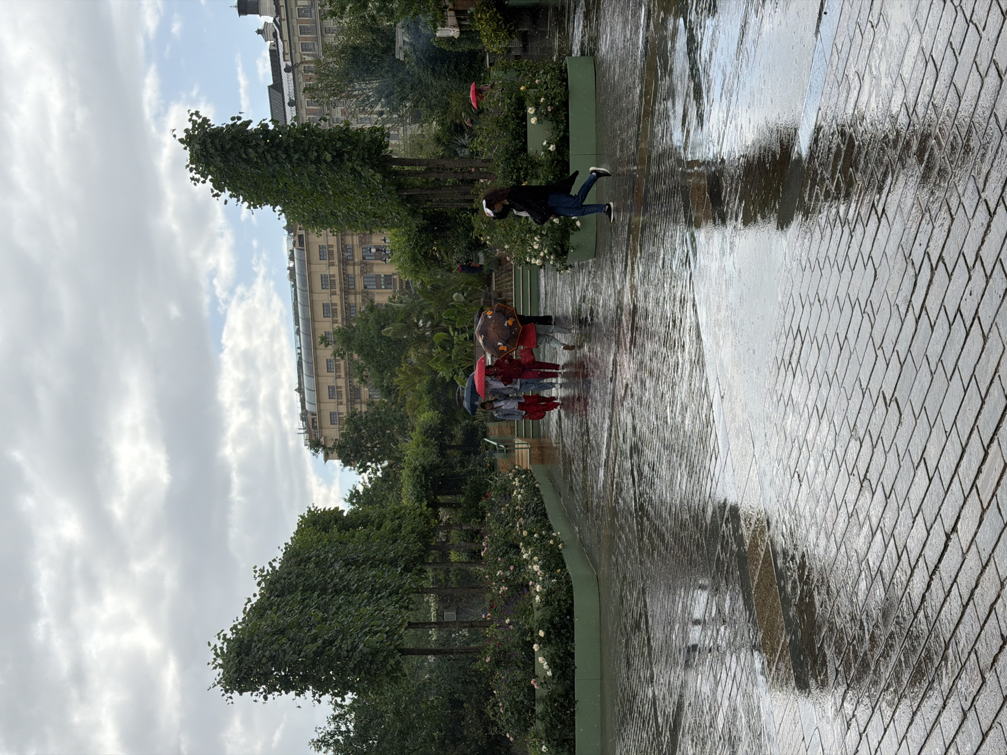 This image depicts a rainy day in a public park area, possibly within a European city given the architectural style of the buildings in the background and the general park design. The ground is paved with rectangular stones and is wet, reflecting the surrounding environment. Several people are visible, most of whom are using umbrellas. One woman in the foreground is walking to the right, her reflection visible in the wet pavement. A group of three people is walking towards the left, also with umbrellas. Trees and flowerbeds line the park area, with a building in the background. The sky is overcast with clouds. The scene appears to be a candid moment captured during inclement weather.