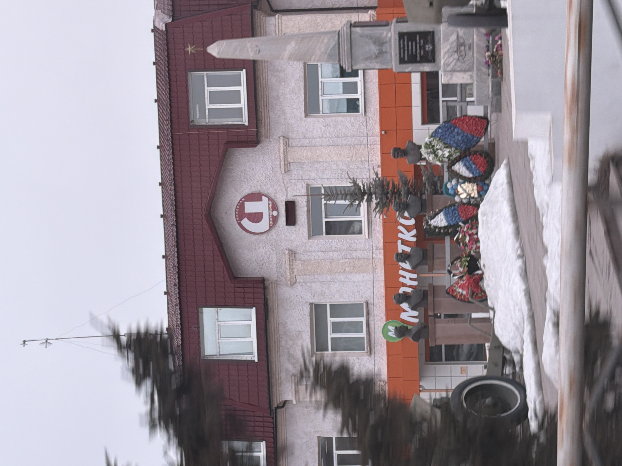The scene depicts a memorial area in front of a light-colored, multi-story building with a dark red mansard roof, located in Beloyarsky, Russia. The building features white-framed windows, decorative facade elements, and a prominent circular sign on its upper facade displaying "ЦЕНТРАЛЬНЫЙ УНИВЕРСАМ" (Central Department Store/Supermarket). Below this, an orange band runs across the ground floor, bearing a green circular logo with an "M" and the partially visible Cyrillic text "МОНЕТКА" (Monetka supermarket). An antenna structure extends from the roof against an overcast sky.
In the foreground, a war memorial occupies the space. It includes a grey stone obelisk topped with a golden star, positioned to the right. To the left of the obelisk, at least three dark-colored busts of individuals are mounted on pedestals. At the base of the busts and around the memorial, several wreaths are laid, predominantly displaying the red, white, and blue colors of the Russian flag, along with some natural flowers. A small evergreen tree stands among the busts. To the far left, a piece of green artillery, possibly a cannon, with a prominent white-rimmed wheel, is partially visible. Patches of snow cover the ground in the immediate vicinity of the memorial. Blurred foreground elements, including parts of trees and a metal railing, suggest the photograph was taken from a moving vehicle or with camera motion. There are no individuals discernible in the image.