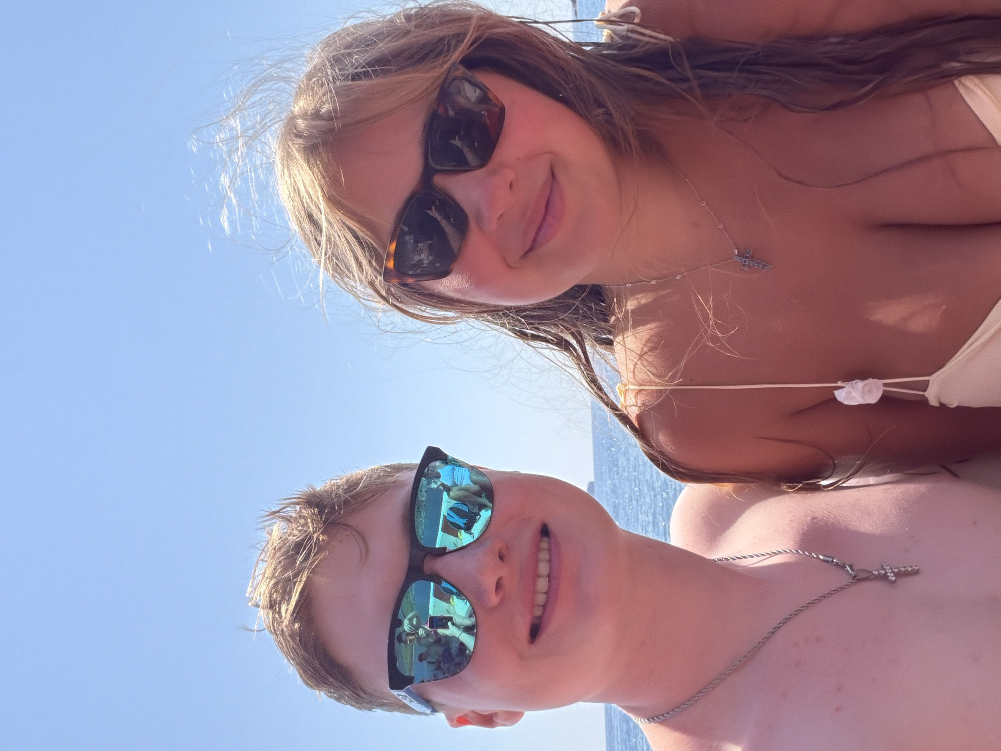 The image is a selfie taken outdoors on a sunny day with a clear blue sky and the ocean in the background. Two young people, a male on the left and a female on the right, are close together, looking towards the camera. Both are wearing sunglasses. The male has fair skin and short blonde hair, and is wearing a silver necklace with a cross pendant. The female has fair skin and long blonde hair, and is wearing a necklace. The reflection in the male's sunglasses shows other people on a boat. The scene appears to be in the city of Kemer, Turkey, suggesting a coastal or maritime environment.